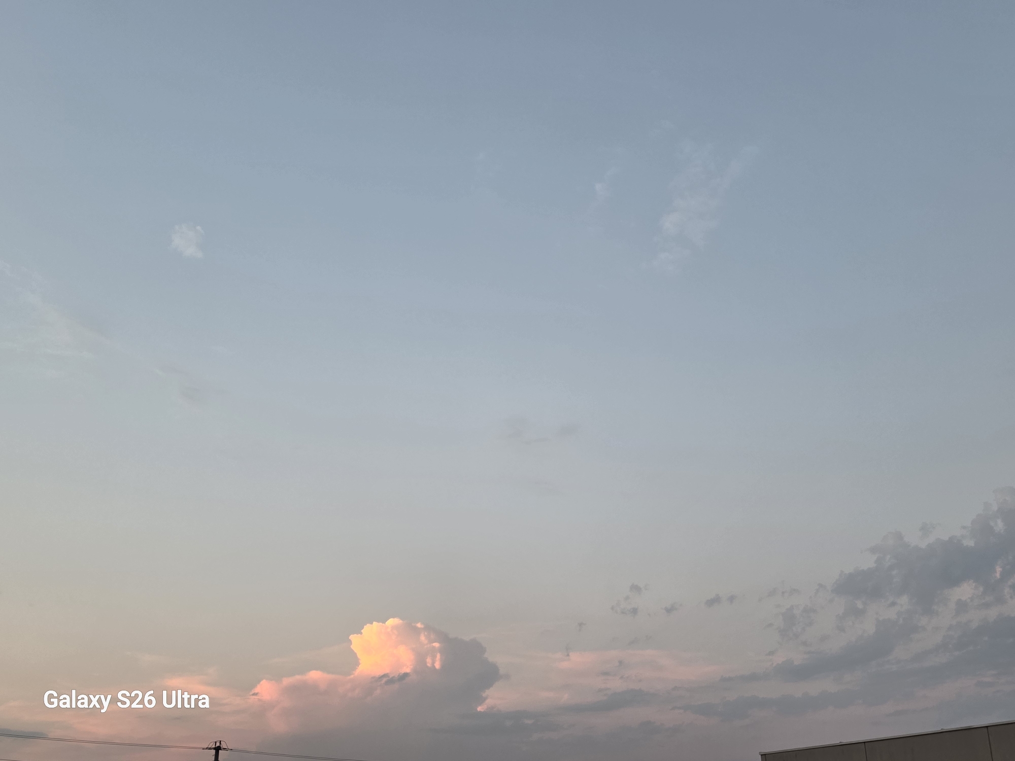 The image shows a clear sky with scattered clouds, tinged with pink and orange hues from the setting sun. The bottom of the frame reveals the top edge of a building, and a power line pole. In the lower left corner, the text "Galaxy S26 Ultra" is visible. This image does not contain any information that would allow for the identification of its location within Amstetten, Austria.