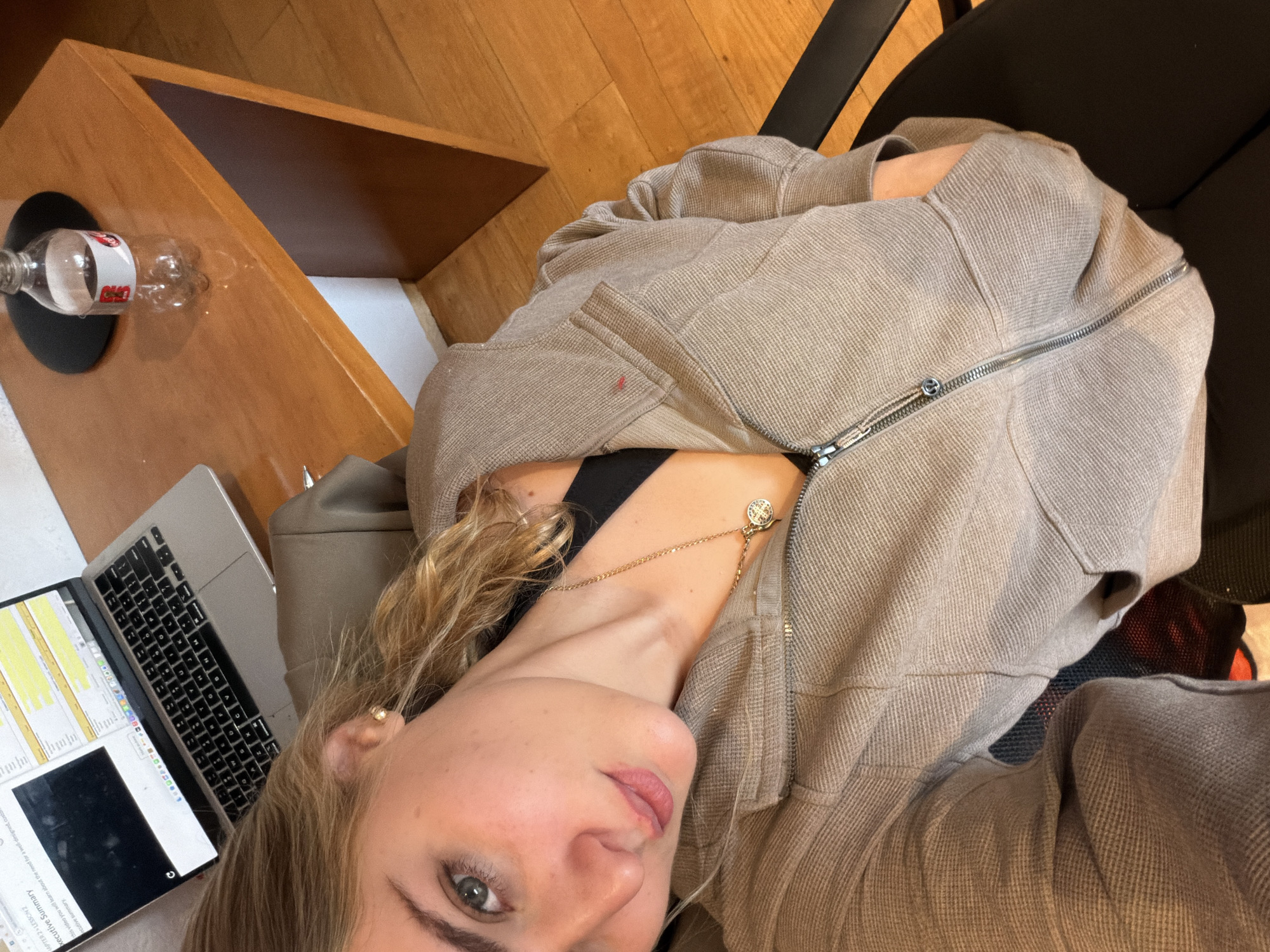 An indoor scene, identified as being located in San Bernardino Tlaxcalancingo, Mexico, depicts a young woman taking a selfie. The woman has light skin, light-colored eyes, and blonde-light brown hair. She is wearing a light brown, waffle-knit, zippered hoodie partially open over a black top, a gold-colored chain necklace with a circular pendant, and a small gold stud earring in her right ear. Her left hand is resting inside the hoodie's pocket. She is seated on a dark-colored chair. To the upper left, on a light-brown wooden desk, an open silver laptop displays a screen with "Executive Summary" and "CHAPTER 2 > LESSON 2" visible, alongside what appears to be a video player interface and a spreadsheet. To the upper right of the desk, a clear plastic Coca-Cola bottle, partially filled with a dark liquid, rests on a black circular coaster. The floor throughout the visible area is light-colored wood.