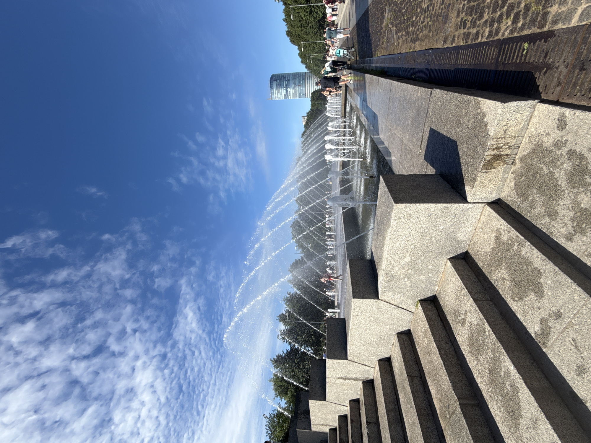This is a ground-level, eye-level, full shot of a large, modern water fountain in a city park on a sunny day with a clear blue sky and scattered white clouds. The fountain is made of granite and features multiple jets of water shooting upwards in arcs. In the foreground, there are wide, tiered granite steps leading up to the fountain area.
To the left of the fountain, a section of the tiered granite structure is visible. To the right of the fountain, a paved walkway runs alongside, bordered by a metal grate. A few people are scattered around the fountain, some walking, and some standing near the water. Behind the fountain, there are trees and a tall, modern glass skyscraper in the distance.
This scene is likely in the area of the Fountain Square (Ploshchad Fontanov) in Saint Petersburg, Russia, which is known for its large, modern fountains and public spaces.