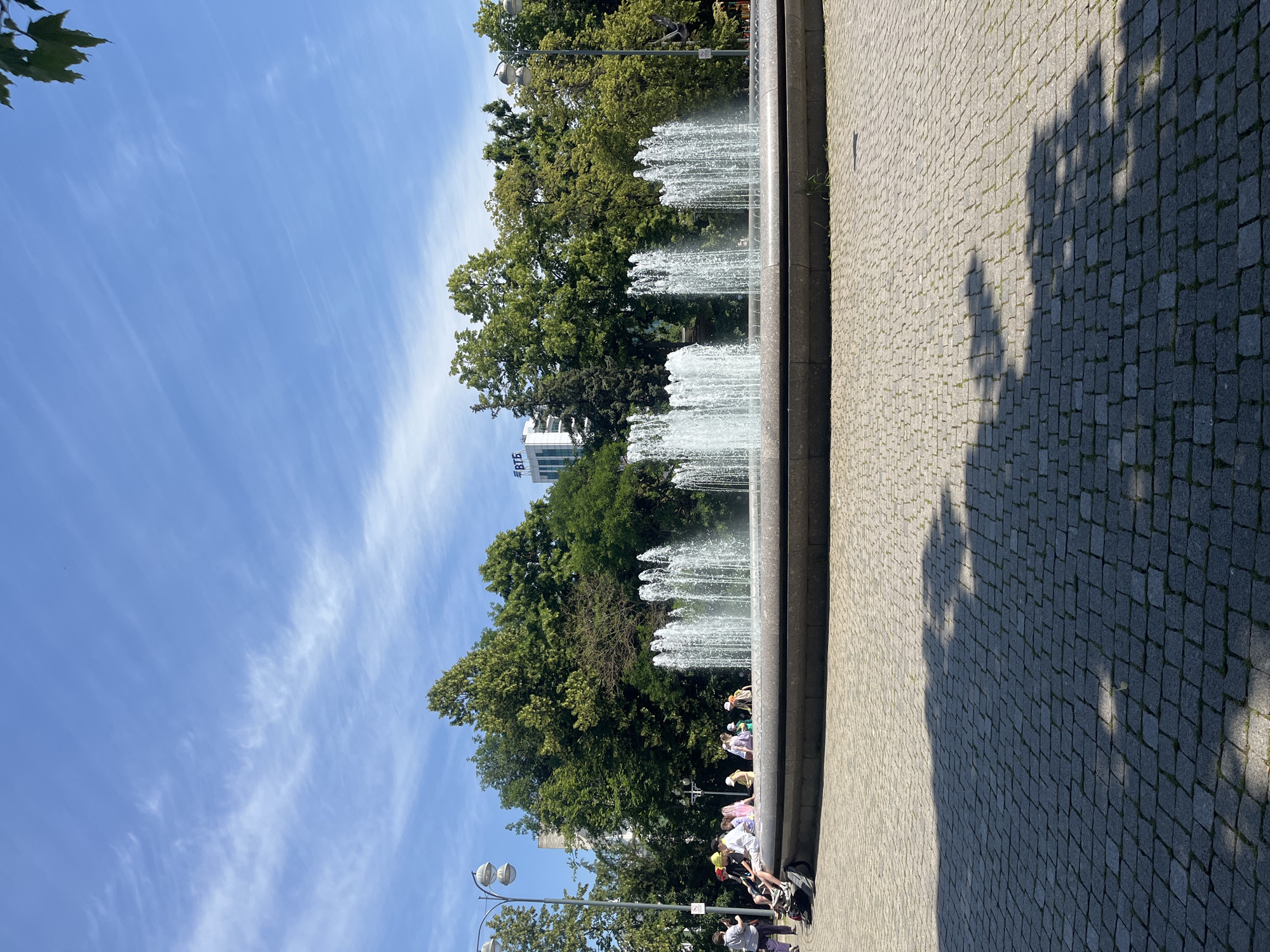 The image depicts a sunny day in a public square, featuring a large, multi-tiered fountain as its centerpiece. The fountain's water streams upwards and outwards, creating a dynamic visual element. Several people are seated on the edge of the fountain, appearing relaxed and enjoying the weather. In the background, a dense line of mature trees forms a verdant backdrop, with a modern white building bearing the Cyrillic inscription "ВТБ" (VTB) visible above the treeline. The ground is paved with grey cobblestones, and shadows from trees are cast across the pavement. The sky above is a clear blue with wispy white clouds. The scene is consistent with the fountain located at Teatralnaya Square in Krasnodar, Russia, a well-known landmark in the city.