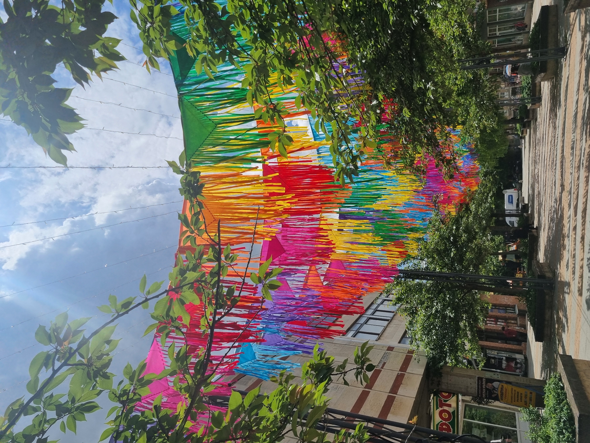 The image captures a street scene in Gorna Oryahovitsa, Bulgaria. Overhead, a vibrant, colorful installation of layered fringed fabric in a spectrum of rainbow hues (green, yellow, orange, red, magenta, purple, and blue) stretches along the street. The installation is suspended by thin cables connected to buildings on either side of the street.
Green trees with dense foliage line the street, partially obscuring the view of the buildings and sky. The sky is visible through gaps in the fabric and trees, displaying a mix of clouds and blue patches. Buildings of various architectural styles are partially visible along the street. At least one van is parked along the street.
The foreground includes foliage from trees, adding a frame to the scene. A pedestrian area is visible on the right.