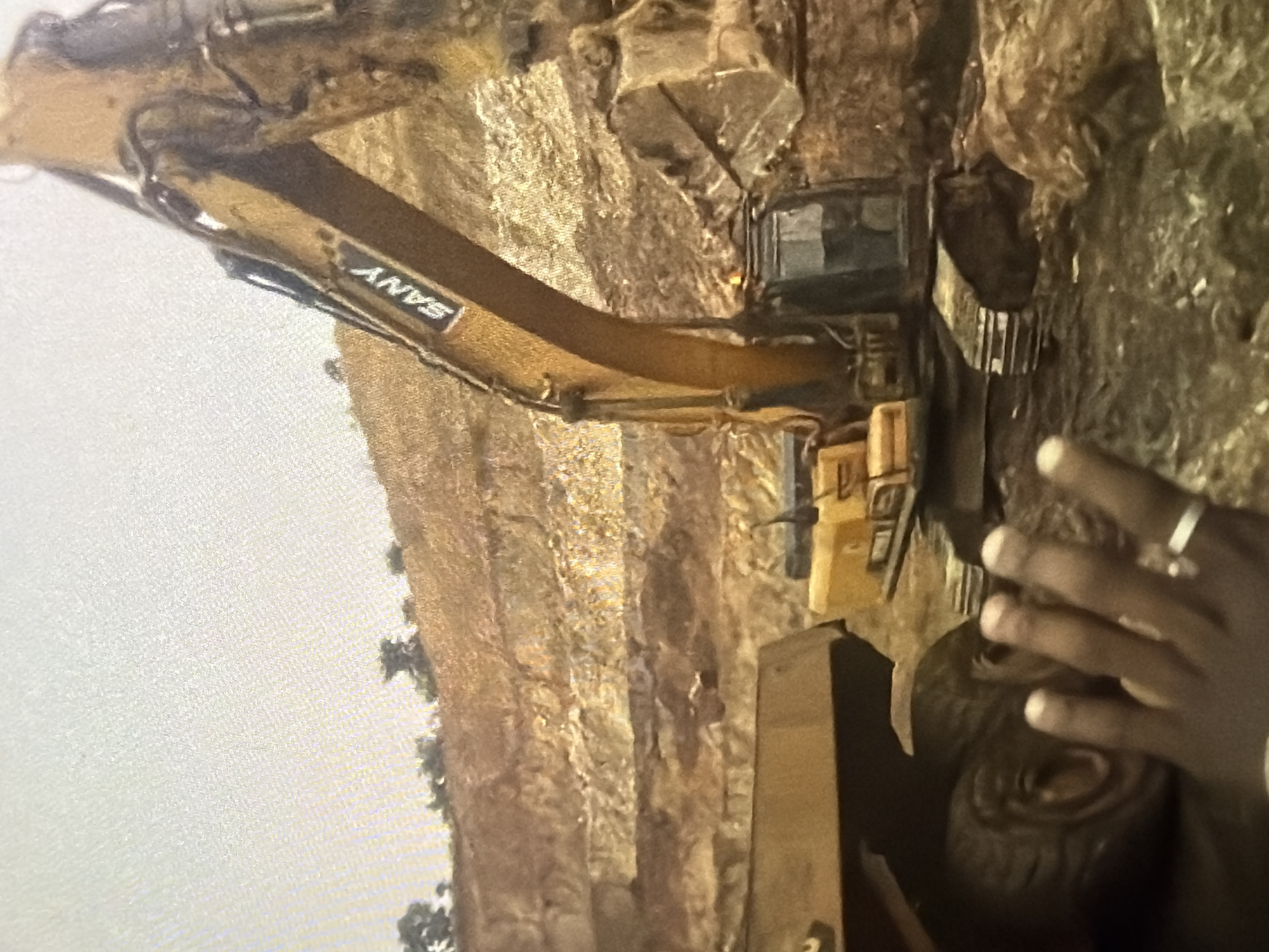The image captures a close-up view of a large yellow excavator with the brand name "SANY" visible on its arm, positioned in a quarry. In the foreground, partially obscuring the scene, is a hand with a ring on one finger. To the left of the excavator, a portion of a large yellow dump truck is visible, including its tire. The background consists of layered, rough-hewn rock faces and a barren, dusty ground. The overall environment suggests an active mining or construction site.