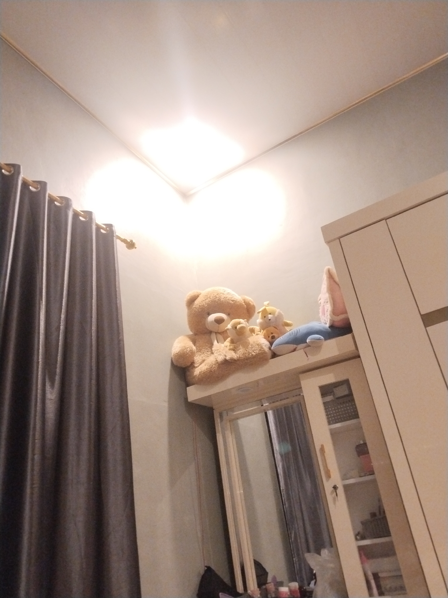 The interior of a room in Kutaglumpang, Indonesia, is depicted in a slightly upward-angled shot. Gray curtains hang vertically on the left side of the frame. A light-colored wall separates the curtains from a tall cabinet or wardrobe on the right. A shelf atop the cabinet holds a collection of stuffed animals, including a teddy bear. The cabinet has a mirrored door and a glass-fronted section with shelves visible inside. The room is illuminated by a bright light source in the ceiling. Personal belongings are partially visible on a surface below the cabinet.
