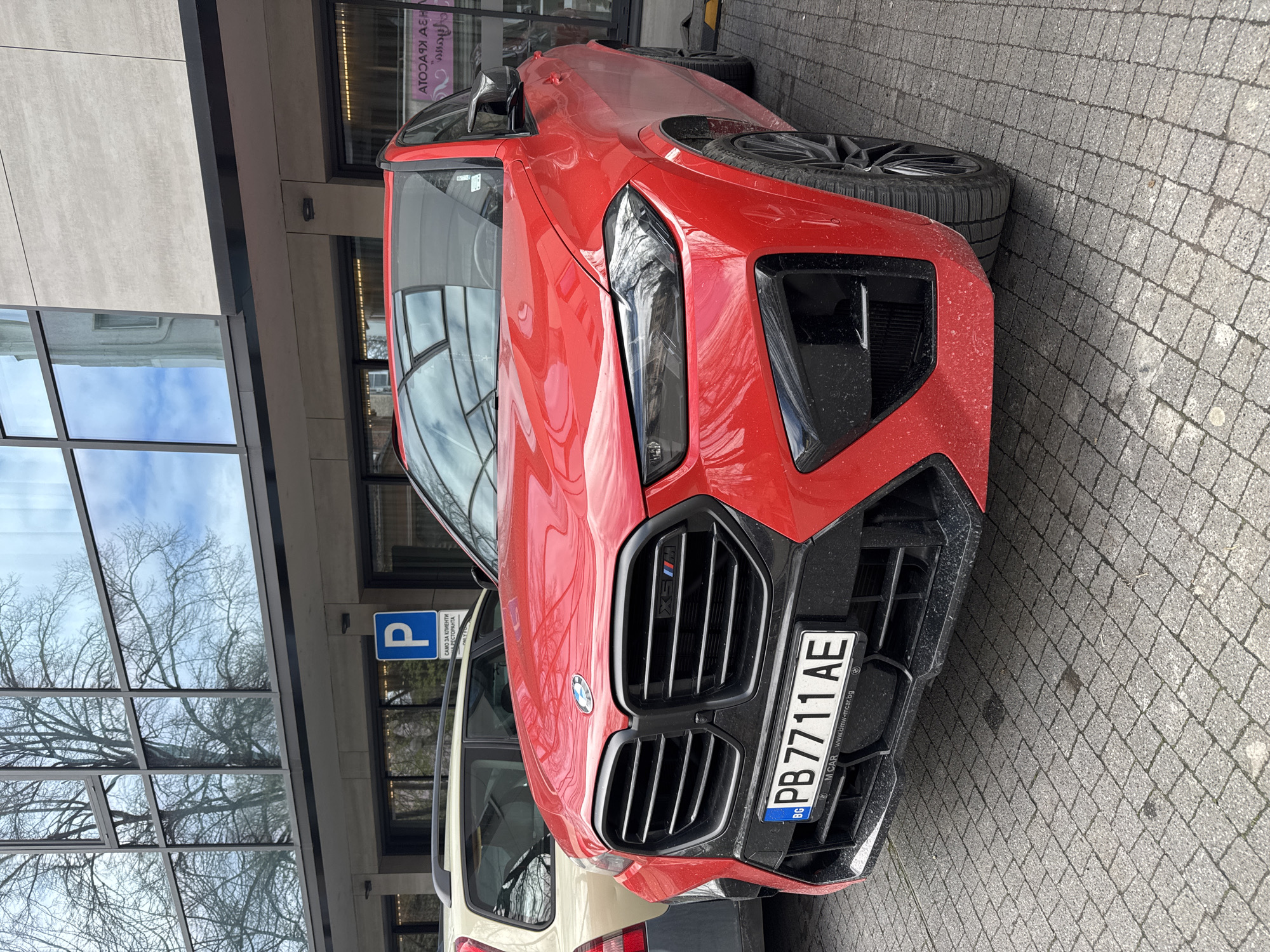 A red BMW X6 M Competition is parked in Plovdiv, Bulgaria. The car has a license plate number of "PB7711 AE". The vehicle is parked on a brick pavement near a building with a parking sign visible. Other vehicles are also parked nearby.