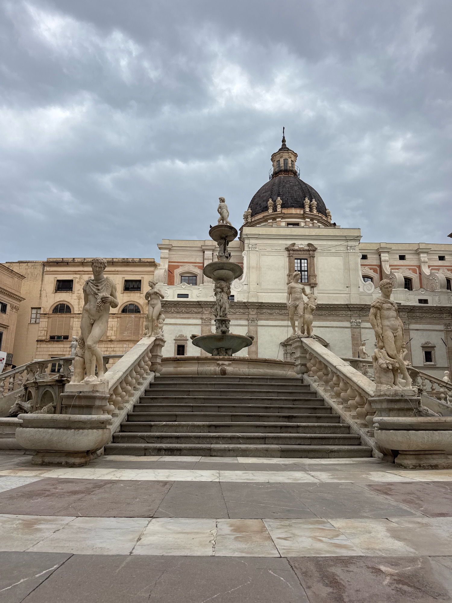 The image shows a wide, ornate stone staircase leading up to a building with a dome. The staircase is flanked by marble statues of male and female figures. The statues appear to be nude and are in classical poses. At the top of the staircase, a fountain is visible with multiple tiers. Behind the staircase and fountain, the building features classical architecture with columns and arched windows. The sky above is overcast and gray. This is the Fontana Pretoria, also known as the Fountain of Shame, located in Piazza Pretoria in Palermo, Italy.