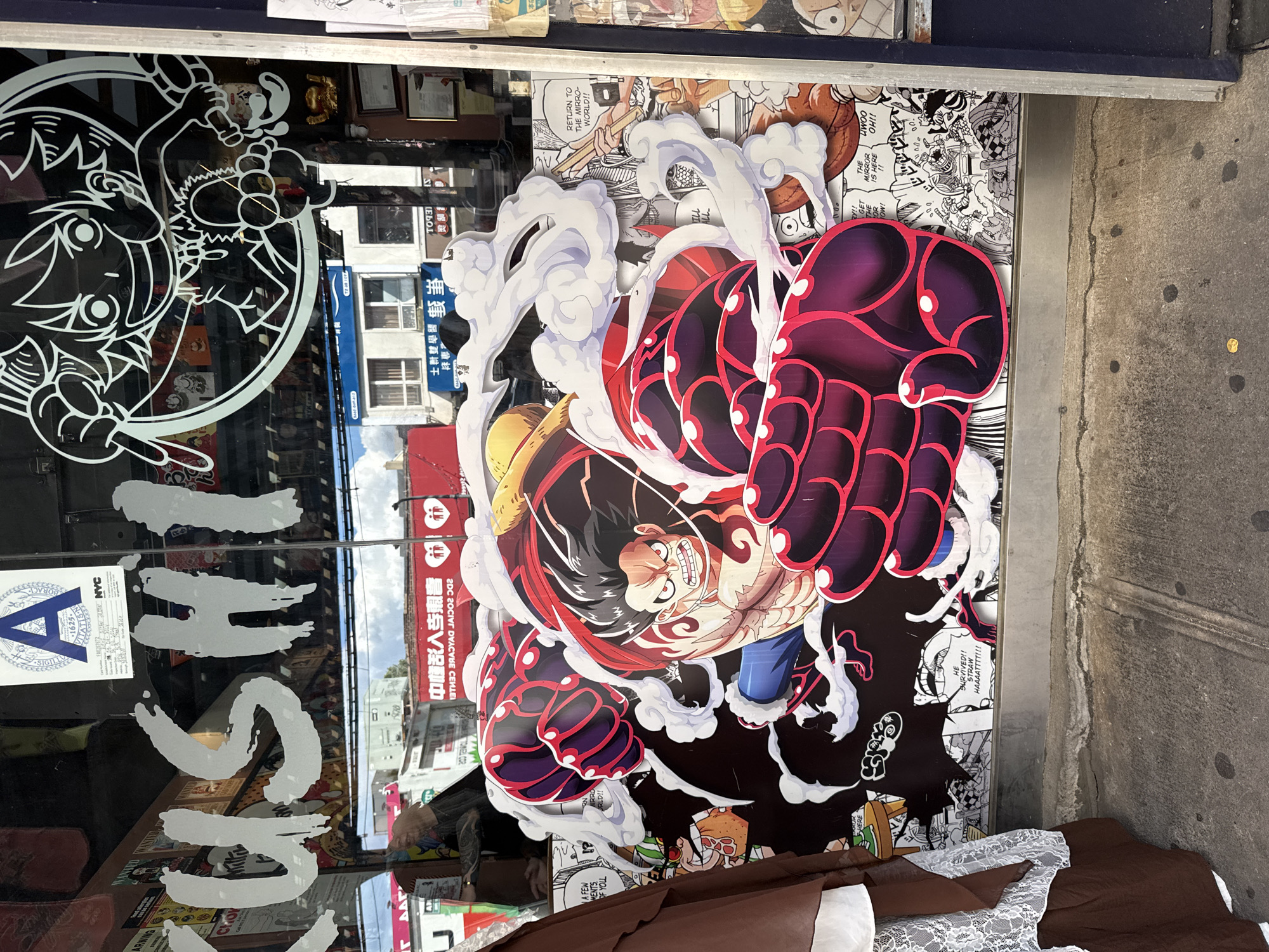 A street-level view of a storefront in New York City, United States of America, features a large graphic display of an anime character from "One Piece" on its glass window. The character, depicted in a dynamic pose with red and purple muscular arms and a determined facial expression, is surrounded by white smoke and comic book panels. Above the graphic, the word "SUSHI" is stenciled in white lettering on the glass, indicating the type of establishment. The storefront appears to be in a commercial area, with other buildings and a street visible in the reflection of the glass. A portion of a mannequin draped in brown and white fabric stands to the left of the display, partially obscuring the window. The ground in front of the store is a concrete sidewalk.