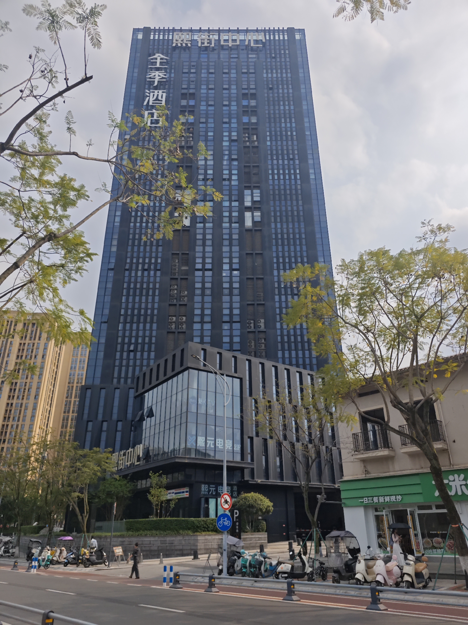 The photo captures a street view in Chongqing High-tech Zone, China. A tall, modern, dark-colored skyscraper dominates the scene, with the words "全季酒店" (All Seasons Hotel) vertically displayed on its side. The building's architecture features a grid-like pattern of windows. At its base, a smaller structure with glass windows and "熙元电竞" (Xi Yuan E-Sports) written on it extends from the skyscraper.
In the foreground, the street is lined with trees, some in partial bloom with yellow-green leaves. Several parked scooters and motorcycles are visible along the sidewalk, indicating urban activity. A few pedestrians are walking along the street, and in the background, a building with a green and white storefront reads "一日三餐新鲜现炒" (Three Meals a Day Freshly Stir-Fried). Signage indicating "P" (Parking) and a bike lane is present.