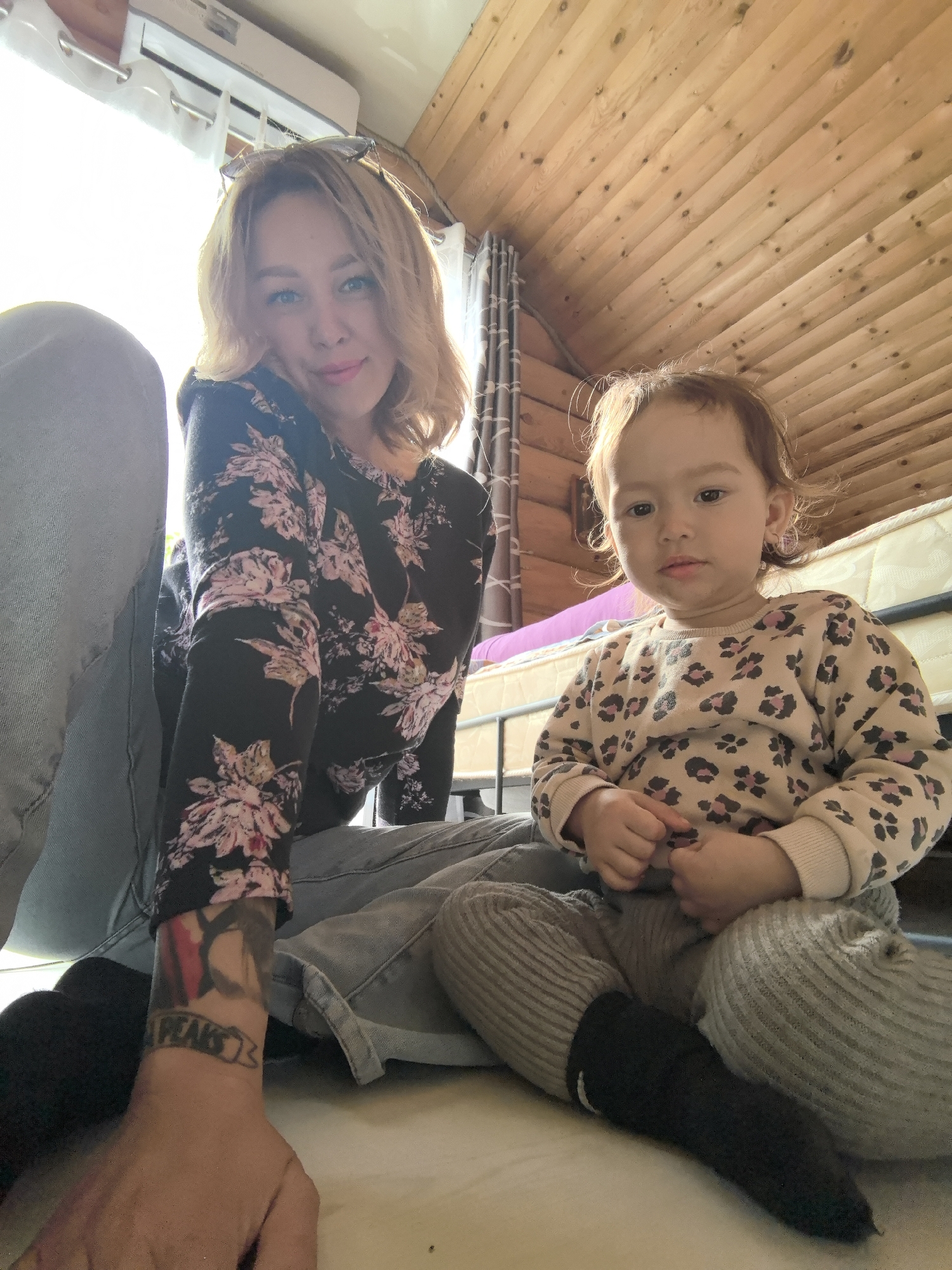A fair-skinned woman with blonde hair and a floral-patterned top sits on the floor, her left arm visible with a tattoo, next to a young child with reddish-brown hair and a leopard-print sweater. The child is sitting cross-legged. They are indoors, under a wooden ceiling. A mattress and curtain are visible in the background. The image was taken from a low angle. The location is likely Noviy, Russia.