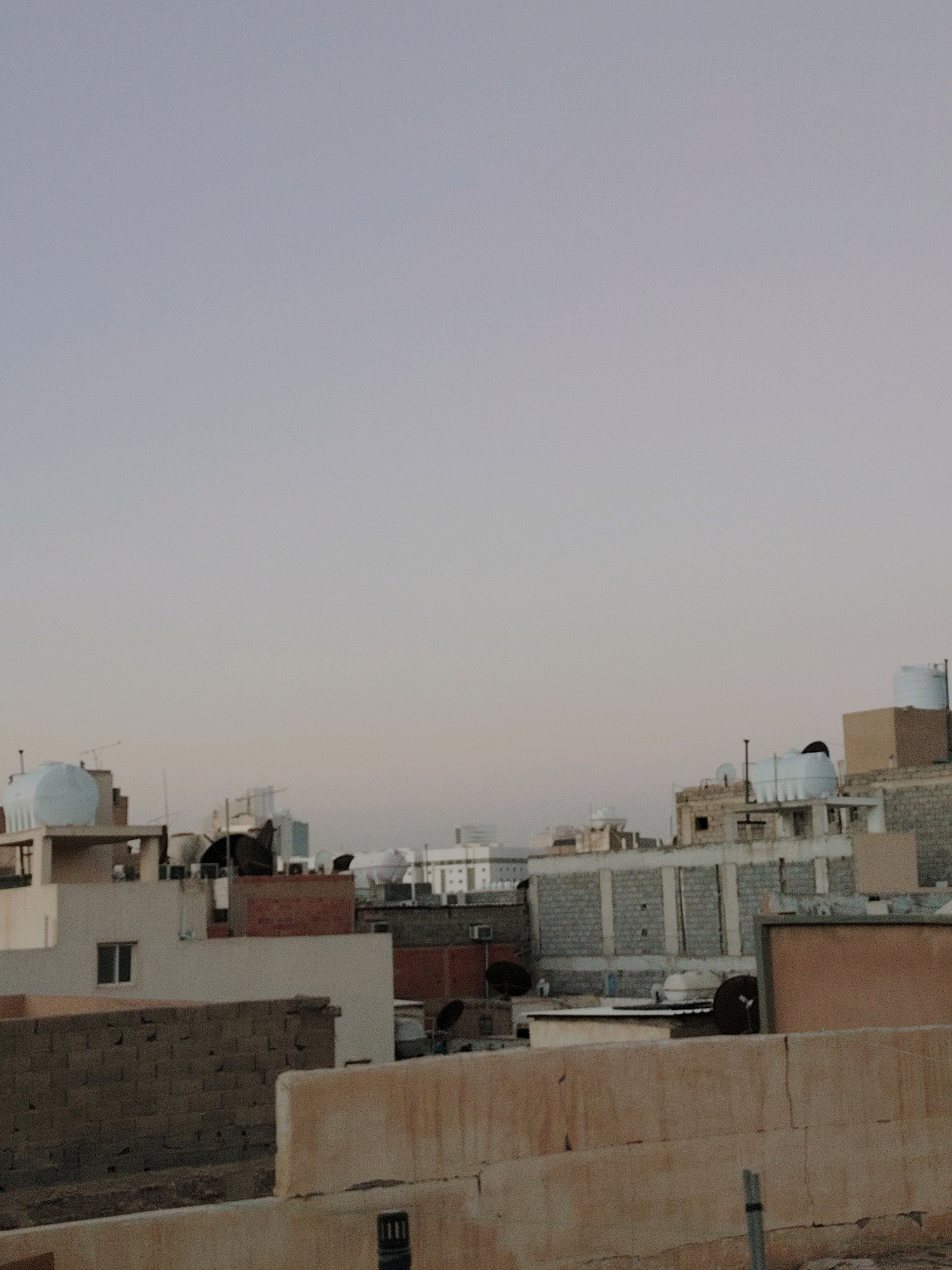 The image depicts a rooftop view of an urban area, likely in Riyadh, Saudi Arabia, given the characteristic architecture and building materials such as concrete blocks and water tanks. The scene is dominated by a muted, overcast sky, suggesting early morning or late afternoon light. Numerous buildings of varying heights and designs are visible, predominantly residential structures with flat roofs. Many roofs are equipped with large, white plastic water storage tanks, a common feature in the region. Some buildings are constructed from lighter-colored concrete or painted white, while others are made of exposed grey concrete blocks or have brick accents. Satellite dishes are also present on several rooftops. In the foreground, a concrete wall partially obscures the view, with a visible metal pipe. The overall impression is one of a densely populated urban environment. There are no people visible in the image.