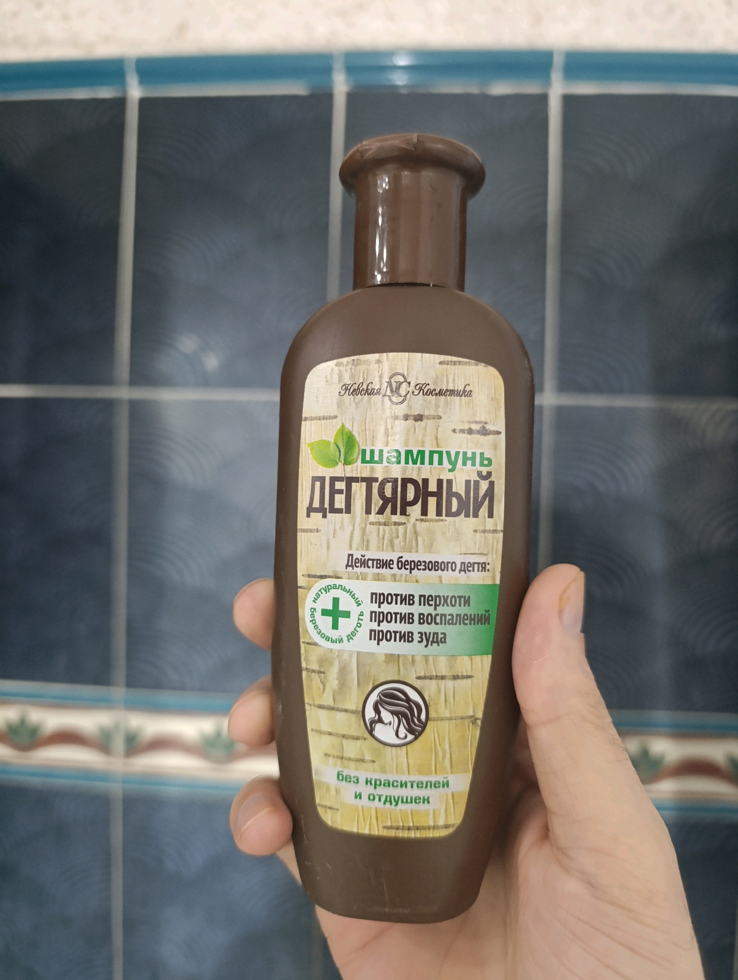 A person's hand is holding a brown shampoo bottle with a light-colored label that features green leaves and text in Russian. The label also includes an image of a woman's head in profile within a circle and text that translates to "for hair health and cleansing." The background is a wall tiled with dark blue squares and thin white grout lines, with a horizontal band of light blue tiles near the top. The lighting suggests an indoor environment.