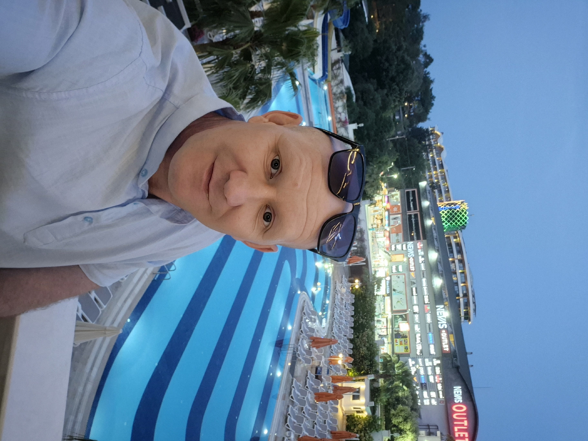 A man in a light-colored shirt and sunglasses stands overlooking a pool with blue stripes, adjacent to the Nevas Outlet shopping center in Alanya, Turkey. The pool is lined with lounge chairs and orange umbrellas. Beyond the shopping center, there is a dark sky. The photo is taken from an elevated position.