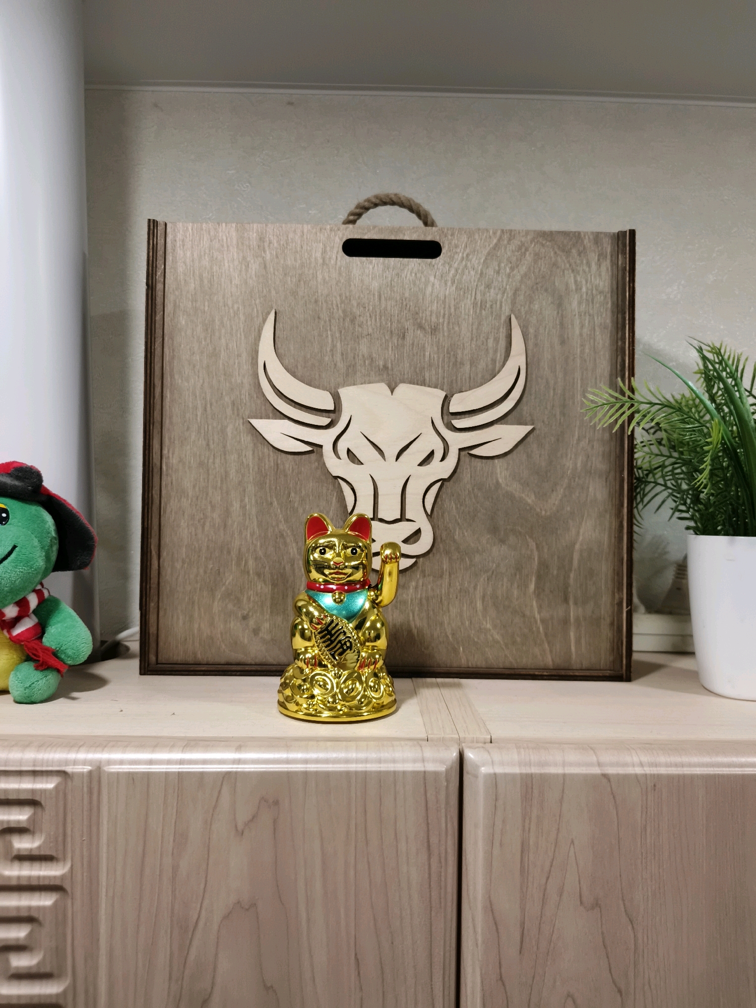 A decorative wooden board with a carved bull's head is placed on a surface. To the left of the board, a green plush toy turtle wearing a red and black hat and a striped scarf is visible. To the right of the board, a golden Maneki-neko (beckoning cat) figurine with a raised paw stands. Behind the Maneki-neko, a potted green plant is partially visible. The scene appears to be indoors, with a light-colored wall in the background and light-colored wooden cabinets or drawers in the foreground. The image is a close-up shot focused on these decorative items, with no indication of the city of Krasnodar, Russia.