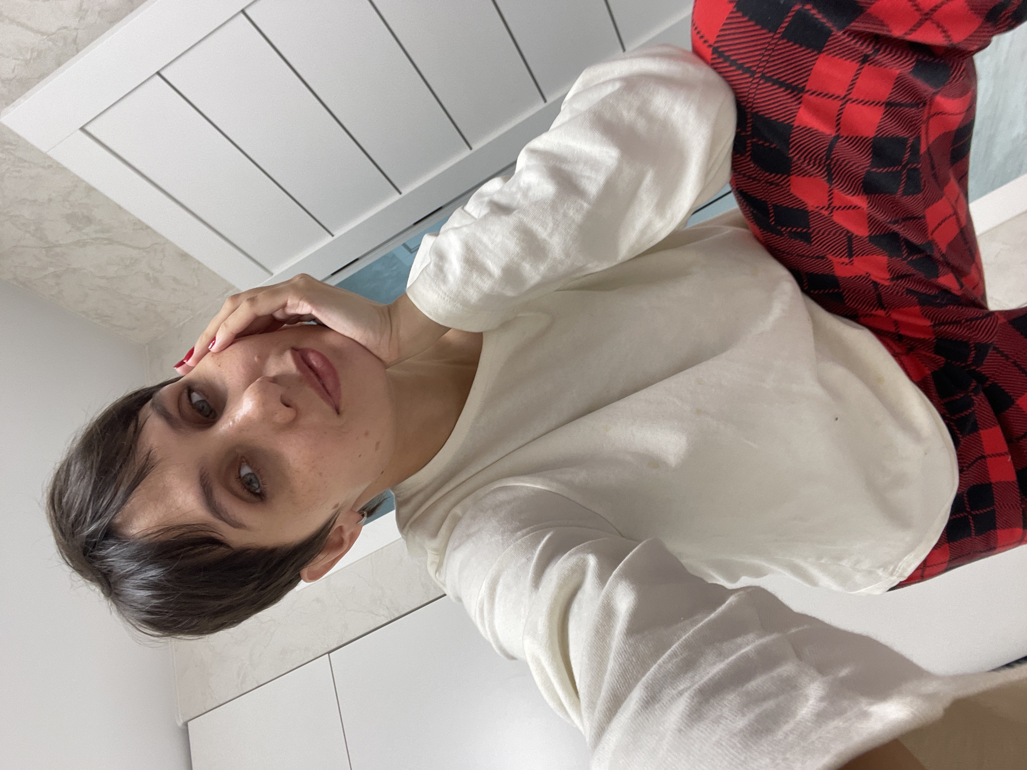 A person with short dark hair, wearing a white long-sleeved shirt and red and black plaid pajama pants, is taking a selfie in what appears to be a bathroom. The background includes white cabinets and a white door with a horizontal panel design. The location, Izhevsk, Russia, is not determinable from the visual information.