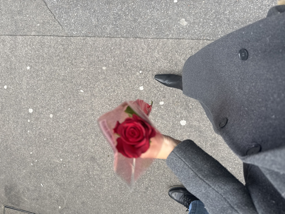 The scene depicts a high-angle view of a sidewalk in Paris, France. A light-skinned human hand, positioned centrally, holds a single red rose encased in clear plastic film. The hand is connected to an arm wearing a dark grey wool coat, visible with two large dark buttons. The right foot of this individual, clad in a black pointed-toe boot, is visible to the lower right. To the lower left, the left foot and lower leg of a second individual are visible, wearing blue jeans and a black lace-up shoe. Both individuals are positioned as if walking forward on the light grey, coarse-grained asphalt or concrete sidewalk, which exhibits scattered small white marks and a faint vertical seam in its texture on the right side.