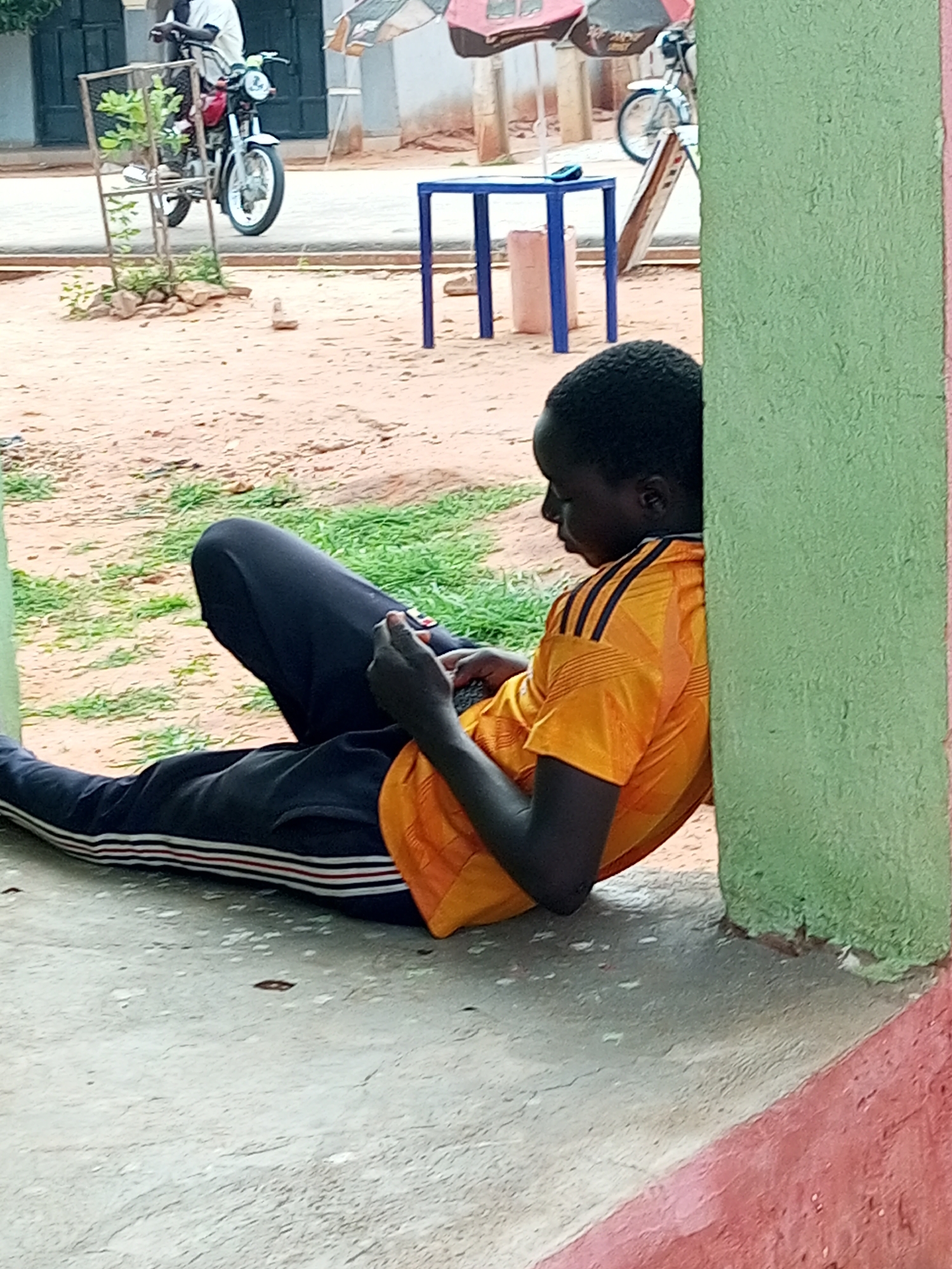 A young boy is sitting on a concrete ledge, leaning against a green-painted pillar, and looking down at a phone in his hands. He is wearing an orange t-shirt with black stripes on the sleeves and dark blue pants with red and white stripes along the sides. His legs are stretched out in front of him. The ground is covered with dirt and some patches of green grass. In the background, there is a motorcycle parked near a metal structure with a plant, and a blue table with a pink object on top. Further back, there are some umbrellas and buildings. The scene appears to be outdoors in a somewhat dusty or sandy area, possibly a community space or marketplace in Tempure, Nigeria.