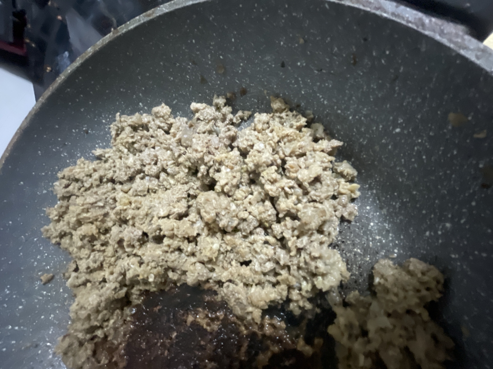 In Bol'shoe Zaborod'e, Russia, a dark-gray, speckled frying pan contains browned ground meat. The meat is clumped together and appears to be cooking or has recently been cooked. Some darker, almost burnt, remnants are visible on the left side of the pan, indicating the meat may have browned unevenly or been cooked for an extended period in that area. The pan is placed on a dark surface, possibly a stovetop, with some white speckled highlights suggesting either reflective surfaces or light glinting off the pan. The background is out of focus, but a dark object with a red element and another dark, shiny surface are faintly discernible.