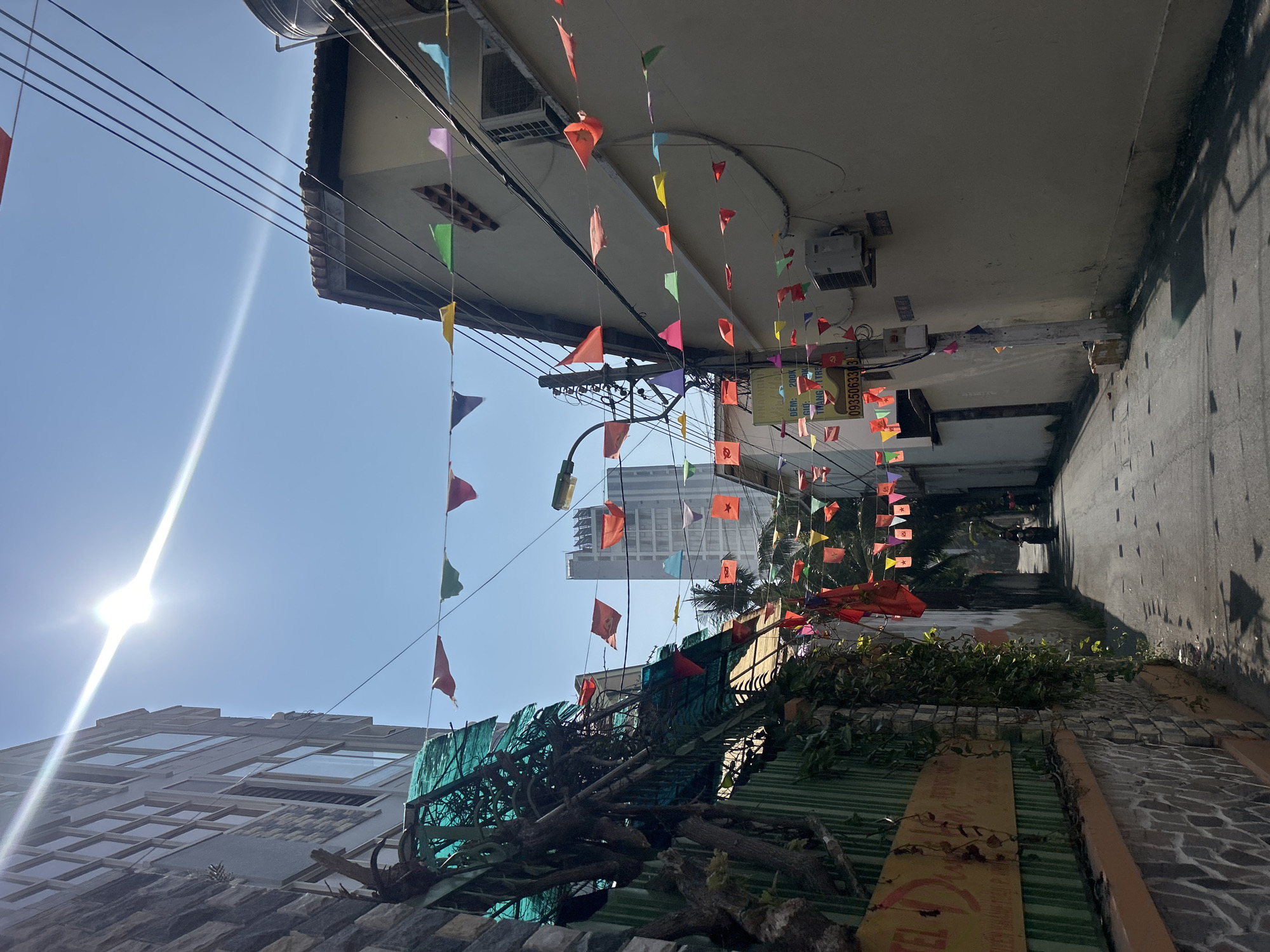The image depicts a narrow alleyway in Bắc Nha Trang, Vietnam. The view is oriented upwards, showcasing the buildings on either side. Colorful triangular flags are strung across the alley, suspended from electrical wires and the buildings' facades. The alley floor is cobbled and merges into an overgrown area with dense foliage and tree branches. A tall building is visible in the distance. Bright sunlight illuminates the sky, creating a stark contrast with the shadows in the alley.