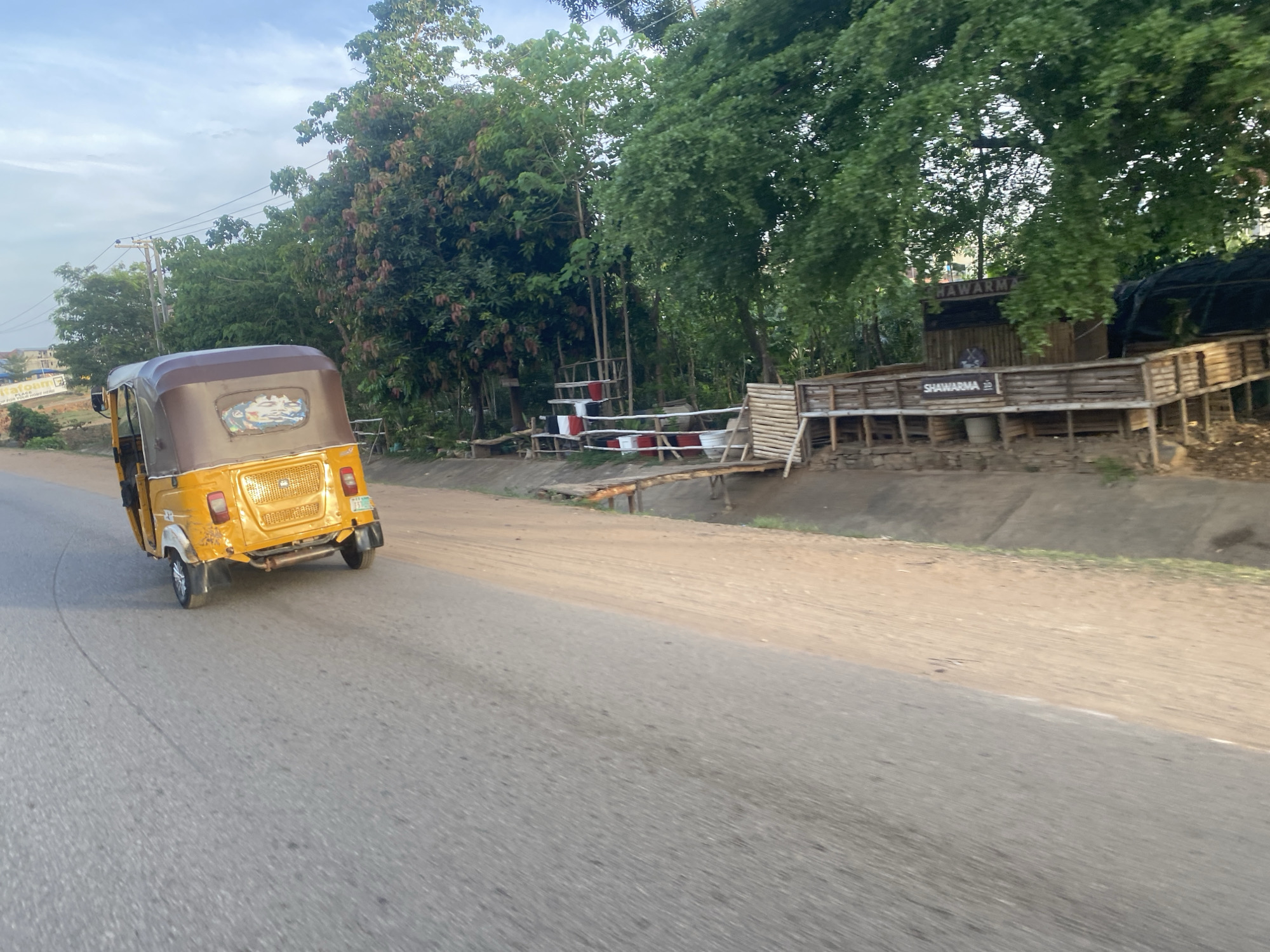 A yellow auto rickshaw travels on a gray asphalt road in Abuja, Nigeria. The rickshaw is moving away from the camera and is on the left side of the road. A row of green trees lines the edge of the road on the right. Behind a fence, a small wooden shack with "SHAWARMA" written on it is seen, suggesting a food vendor. The sky is overcast, with light blue tones. Power lines are visible above the trees. The road has a slight curvature and appears worn.