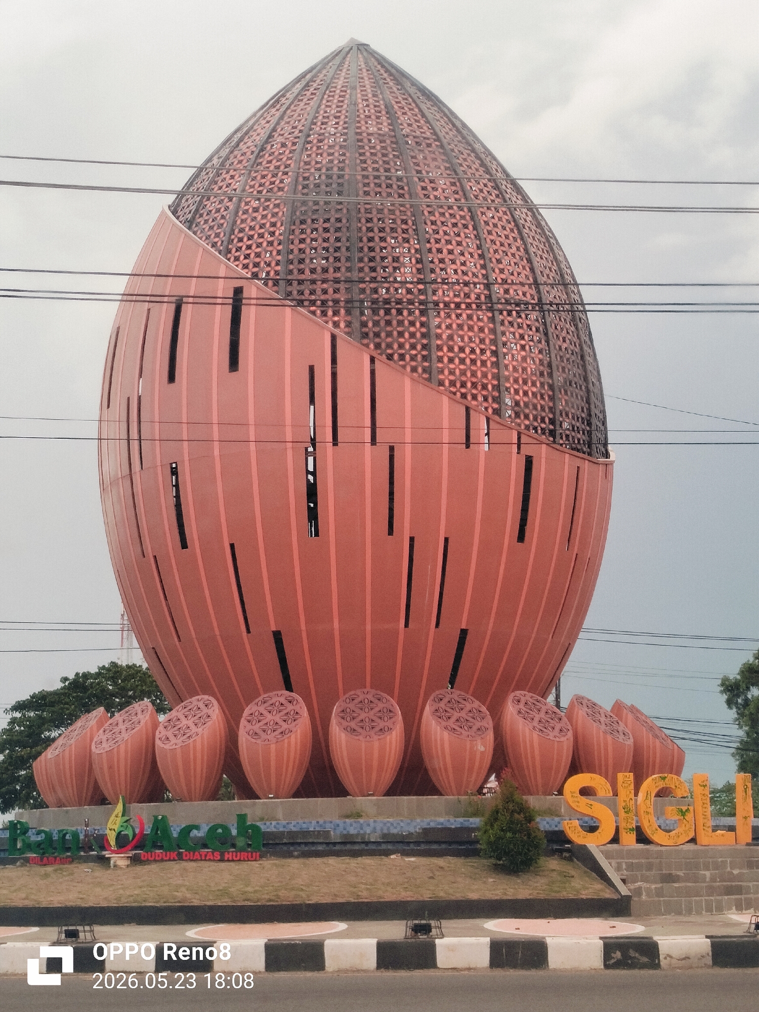 The image depicts a large, monument-like structure in Sigli, Indonesia. The primary structure is an egg-shaped or oval edifice, seemingly made of a reddish-brown material. The upper section has a lattice-like pattern with geometric openings. The lower section has a vertically ribbed surface with narrow, vertical slots. The structure rests on a base adorned with smaller, similarly colored but differently designed ellipsoidal objects. The word "SIGLI" is spelled out in large, yellow block letters on the right side of the structure. In front of the main structure, there is greenery and the words "Bank Aceh," along with what appears to be a logo. The foreground shows a portion of a road with a black-and-white striped border. Overhead power lines are visible against the sky. The background includes trees and an overcast sky. The image is captured from a low angle, emphasizing the monument's scale. A timestamp "OPPO Reno8 2026.05.23 18:08" is visible in the lower left corner.