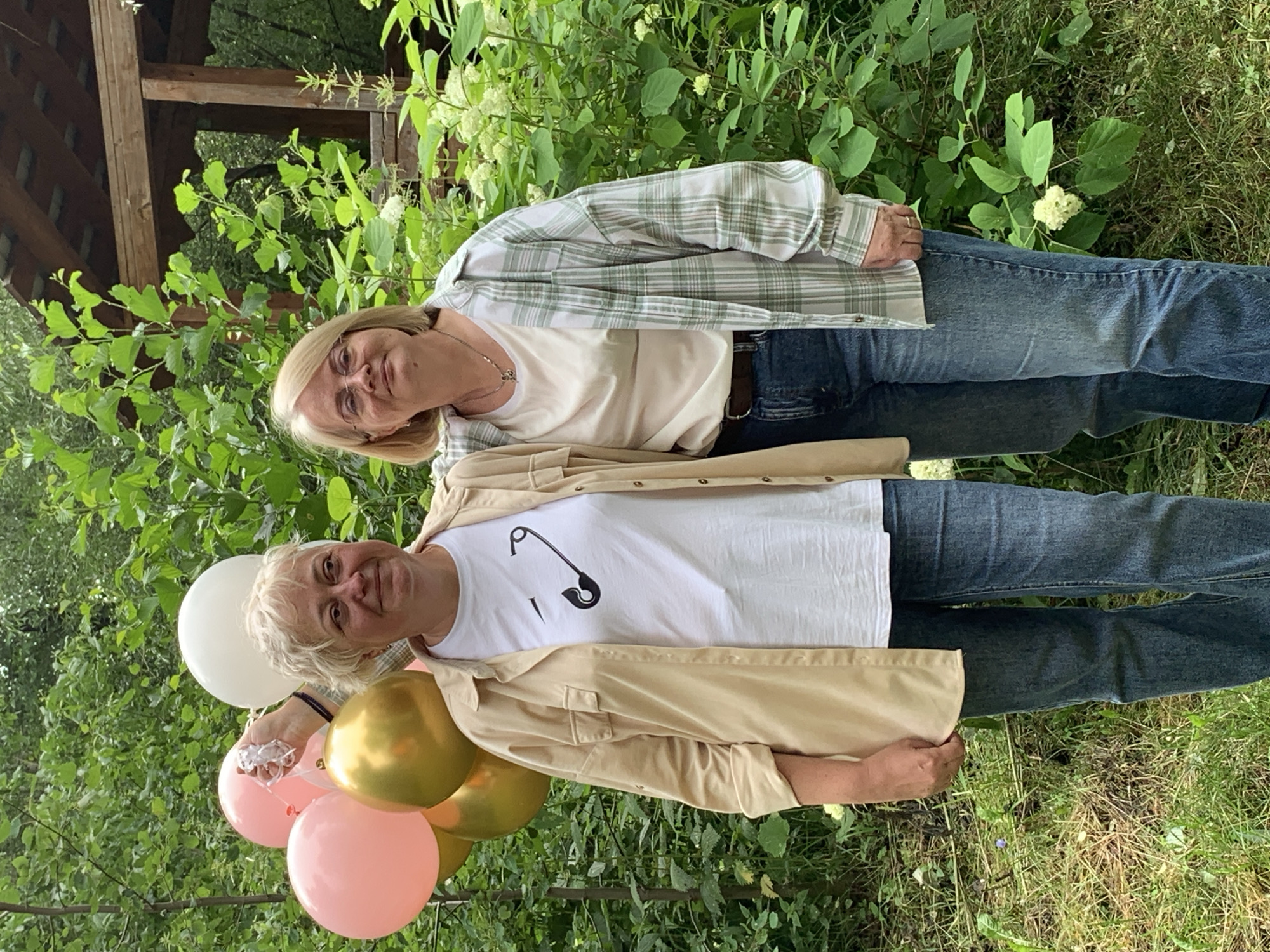 Two women, who appear to be of similar age, are posing for a photo outdoors. They are both wearing casual clothing. The woman in the foreground is wearing a beige open-collar shirt over a white t-shirt with a safety pin graphic, and jeans. She is holding a bunch of balloons, including pink, gold, and white. The woman in the background is wearing a light green and white checkered shirt over a cream-colored t-shirt, and jeans. She is wearing glasses and has blonde hair. The scene is set in a natural environment with lush green foliage and trees. There is a wooden structure, possibly a gazebo or part of a building, visible in the upper left corner of the image. The lighting suggests it is daytime, likely with overcast or diffused sunlight, as there are no harsh shadows. The overall mood of the image appears cheerful and relaxed, possibly celebrating an event given the balloons. The location is identified as Chaikovsky, Russia. There is no visible text in the image.