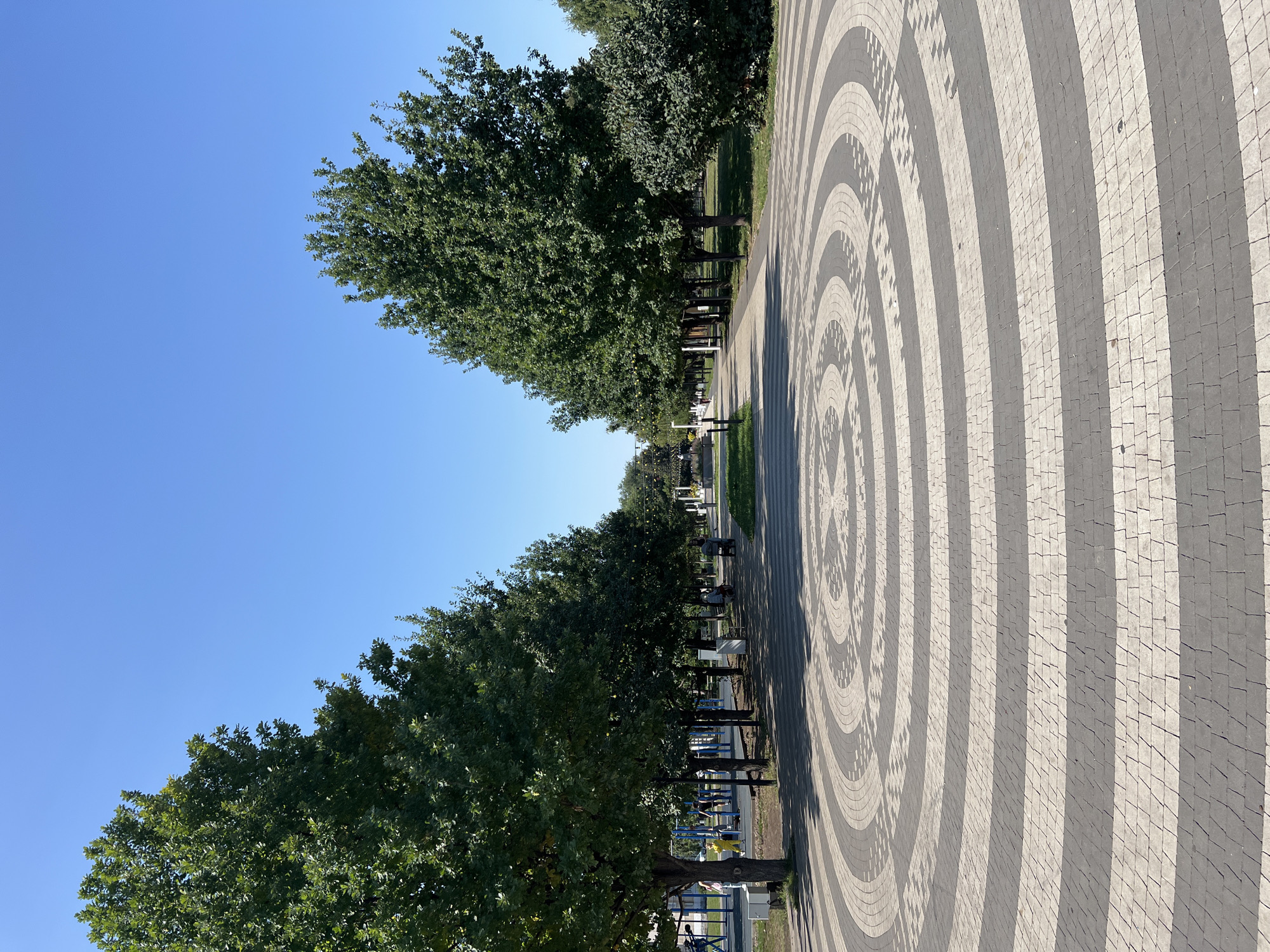 The image depicts a sunlit park in Salavat, Russia, featuring a circular paving stone design with alternating dark grey and light grey concentric rings. Two rows of mature deciduous trees line a paved pathway extending into the distance. The left side of the path borders a playground with blue and yellow structures and several individuals, including a child in a yellow outfit, a woman, and a man. Further down the path, to the right, benches are visible, with a person seated on one and another in a wheelchair to the right of them. The sky is clear and blue.