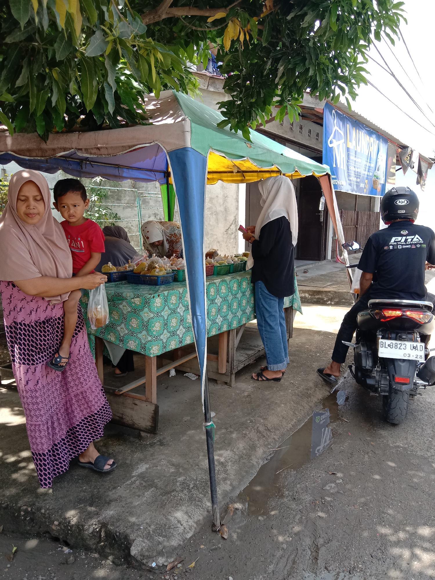 The image depicts a street scene in Lhokseumawe, Indonesia. A vendor stands behind a covered table displaying packaged food items. A woman in a headscarf and a patterned dress holds a young boy in her arms while looking at the food. Another woman, also wearing a headscarf, stands at the table, seemingly making a purchase. A man on a motorcycle, wearing a helmet and a t-shirt with text and a logo, is positioned beside the table. A laundry banner is visible in the background. The road is cracked and wet, with a puddle reflecting the street scene. The motorcycle's license plate reads "BL 3823 UAD 03-28". The scene is partly shaded by a tree.