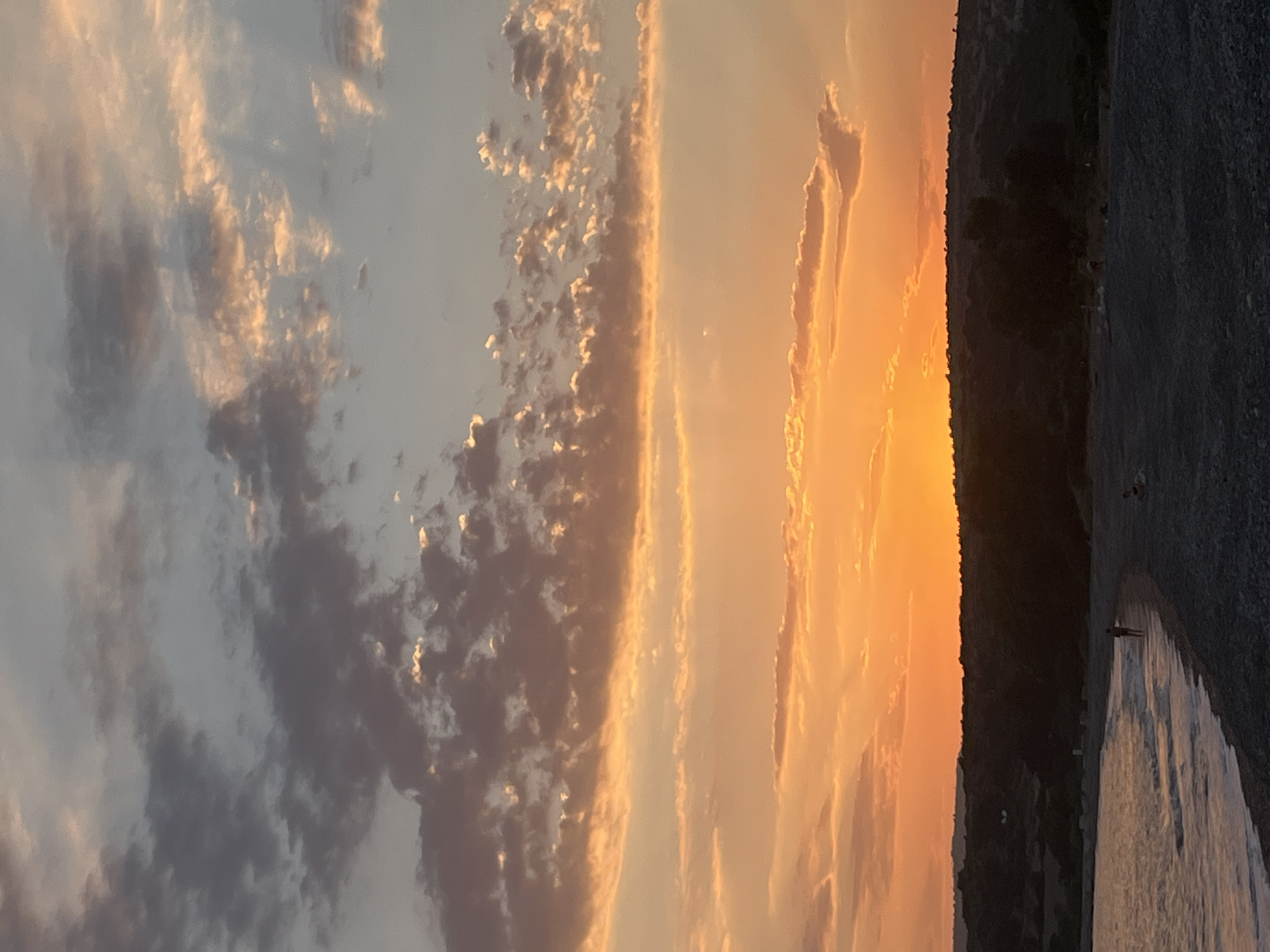 The image depicts a sunset scene at Avdimou beach in the Sovereign Base Areas of Akrotiri and Dhekelia. The sky is ablaze with warm colors, transitioning from bright orange near the horizon to softer hues of pink and gray higher up, with scattered clouds. The sun is just dipping below a dark, silhouetted landmass that forms the backdrop of the beach. The sea is calm, with gentle waves washing onto a pebble-strewn beach. A lone figure is seen walking along the water's edge, and a few other people are scattered on the beach, appearing to relax or observe the sunset. The overall impression is one of tranquility and natural beauty at the end of the day.