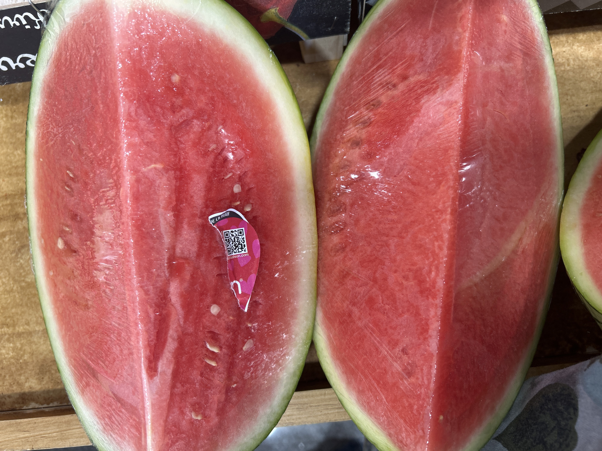 The image shows two halves of a watermelon, cut open and placed side-by-side. The watermelon flesh is a deep red color and appears juicy. Seeds are visible in the left half. A sticker with a QR code is attached to the left half of the watermelon. The watermelon halves are on a wooden surface. The image includes parts of another cut watermelon on the right side.
The QR code on the sticker indicates the watermelon is from "DE LA PRADA fashion.com", which references a Spanish company located in Barcelona, Spain.