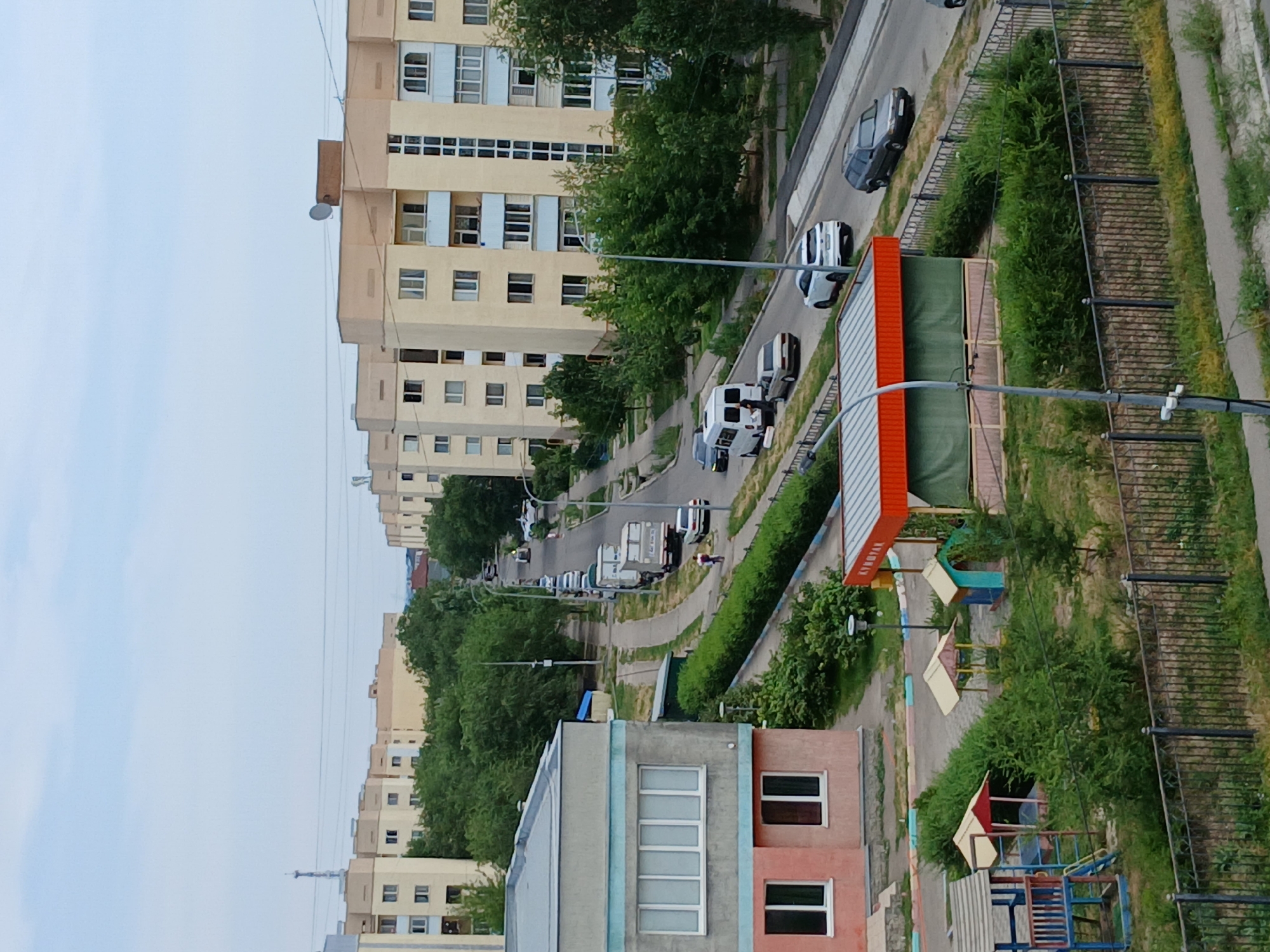 The image depicts a street scene in Almaty, Kazakhstan, with several multi-story apartment buildings lining the road. The buildings are predominantly light yellow with beige trim. A paved road curves through the scene, with several vehicles visible, including cars, a white van, and a truck with a trailer. Trees and green bushes are planted along the sides of the road and in front of the apartment buildings. In the foreground, there is a playground with colorful structures and a covered pavilion with an orange roof. A black metal fence runs along the edge of the playground area. The sky is overcast.
