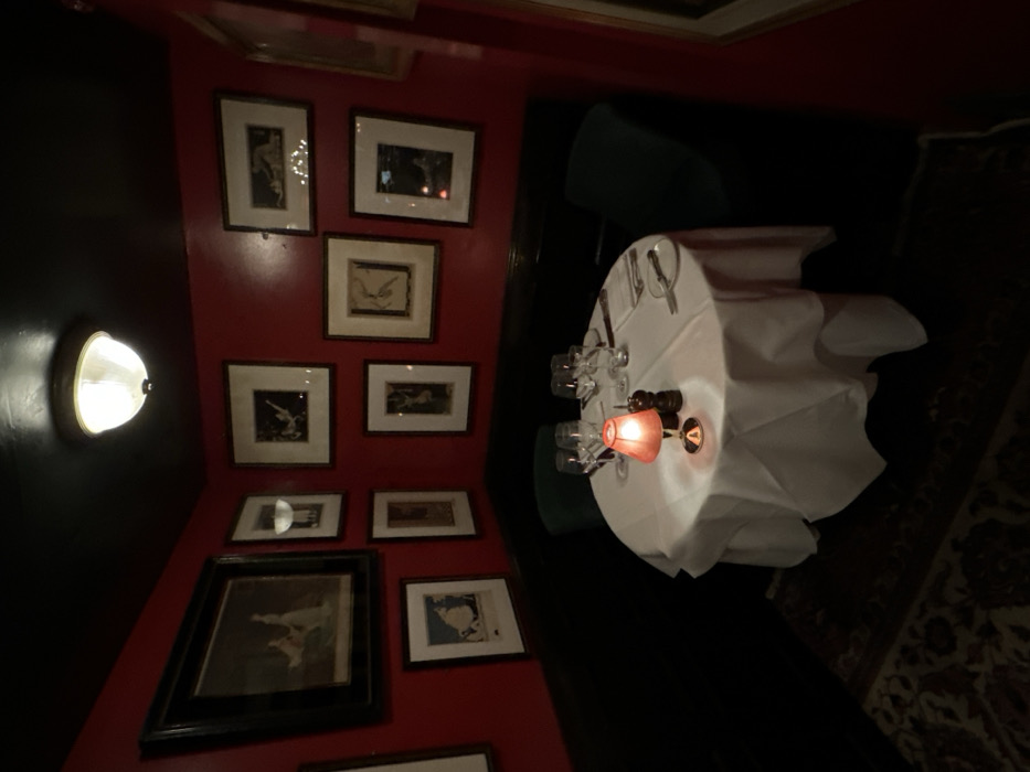A dimly lit, red-walled restaurant booth features a round table draped with a white tablecloth. The table is set for four with glasses, cutlery, and a small lamp emitting a warm glow. Black and white framed pictures adorn the wall, illuminated by a wall sconce. Dark green chairs surround the table. A portion of a patterned rug is visible on the right.