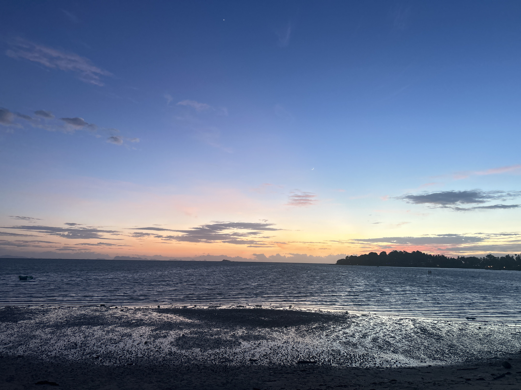 The image captures a serene twilight scene at a beach in the Ko Pha-ngan District of Thailand. The sky transitions from a deep blue at the zenith to soft hues of pink, orange, and yellow near the horizon, where a sliver of the sun is visible. Scattered clouds are present in the sky. The calm sea stretches across the mid-ground, reflecting the colors of the sky and exhibiting subtle wave patterns. In the foreground, the wet sand of the beach is visible, appearing dark and reflective due to the receding tide. The shoreline is dotted with small rocks and debris. On the horizon, a distant landmass, likely an island or the mainland, is silhouetted against the colorful sky, with trees and buildings discernible. A small boat is present on the water to the left of the frame, and two figures can be seen standing in the shallow water on the right side of the image. The overall atmosphere is peaceful and tranquil, suggesting the end of the day.