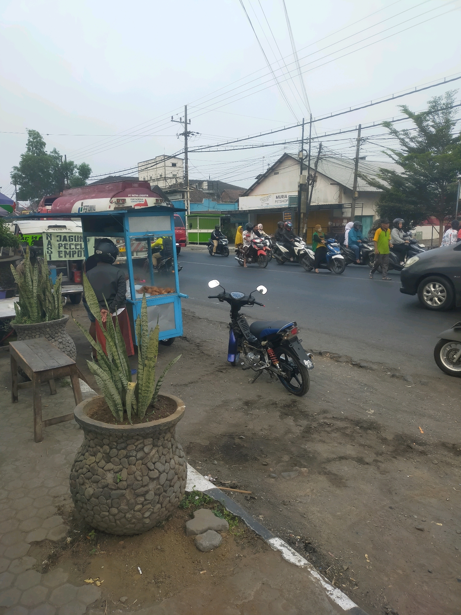 A street scene in Bululawang, Indonesia, features a food cart selling "JAGUNG PECEL EMPO," with a vendor attending to customers. Several motorcycles are parked and in motion on the road, alongside a Pertamina tanker truck and a black car. The environment is urban, with buildings, power lines, and trees visible. Snake plants in large stone pots are placed along the roadside. The sky is overcast.