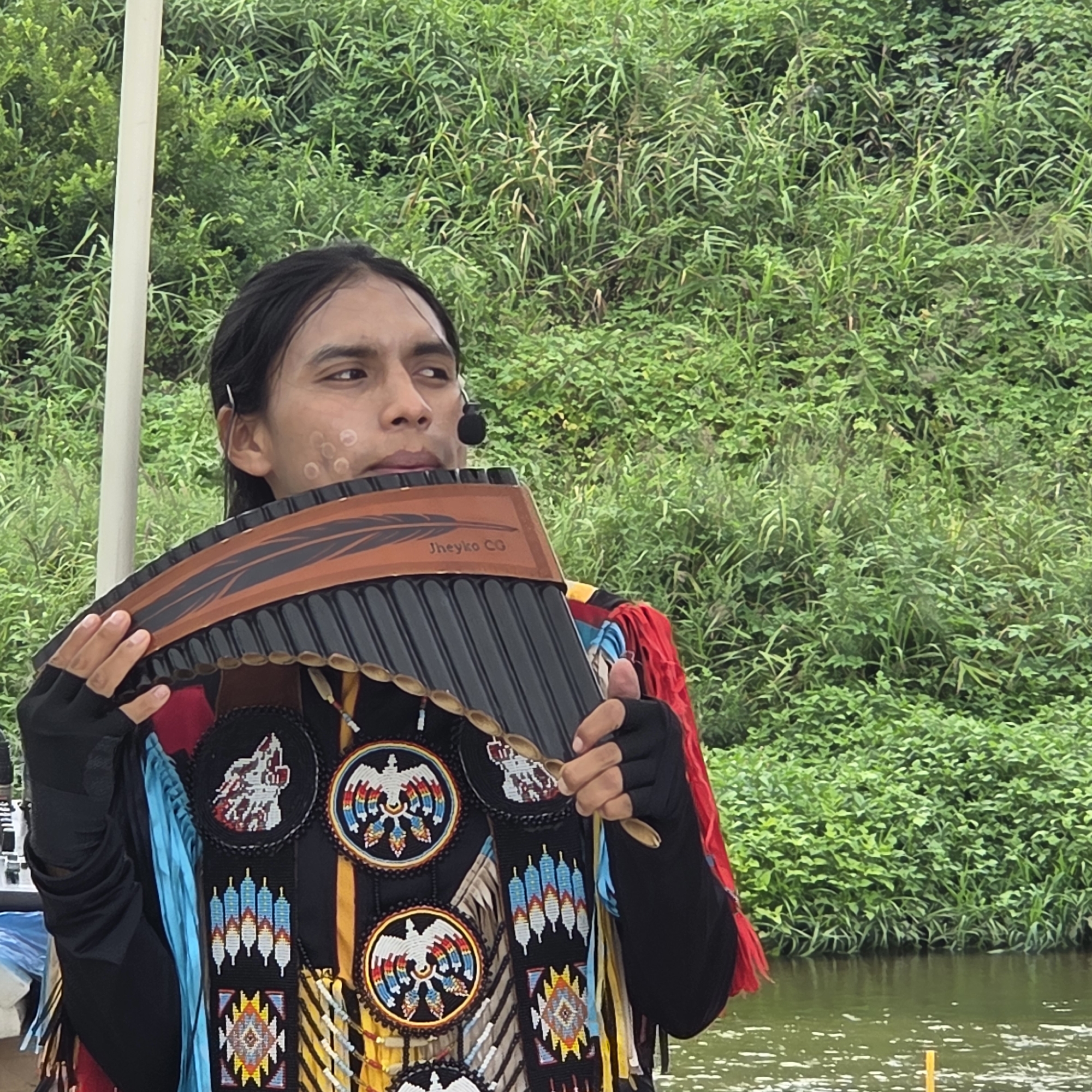 A young man, appearing to be of Indigenous descent, stands outdoors playing a large pan flute. He is adorned in traditional clothing that includes a black shirt with intricate beaded designs depicting eagles, wolves, and geometric patterns. He wears a microphone headset. The pan flute has a brown and black wooden casing with a feather design and text that reads "Jheyko CG". The man is positioned in front of lush green foliage, with a body of water visible in the background. The scene is likely an outdoor cultural event or performance.