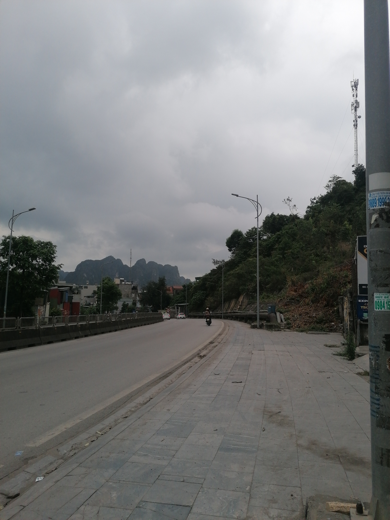 The image depicts a street scene in Ha Tu Ward, Vietnam, on an overcast day. A gray concrete road curves gently to the left, bordered by a concrete barrier and a gray tiled sidewalk. A motorcycle is traveling on the road. In the distance are buildings and a mountain range. A telephone tower is visible on top of a hill to the right of the road. The right side of the sidewalk is overgrown with vegetation. A utility pole is on the right of the frame. The sky is filled with heavy gray clouds.
