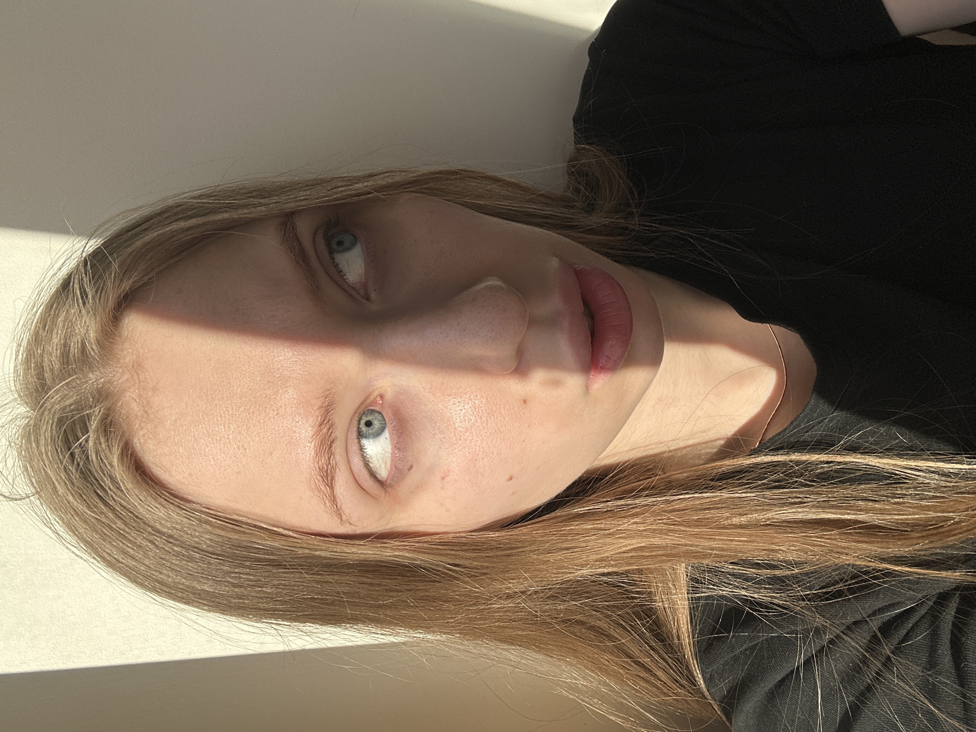 A close-up portrait depicts a young adult female indoors in Chaikovsky, Russia. She has long, light brown hair, blue eyes, and fair skin with visible moles and faint redness. She wears a black t-shirt and a thin, gold-colored choker necklace. Her head is angled slightly, and her gaze is directed upwards and to her right. Her mouth is slightly open, revealing a pinkish hue on her lips. Strong, directional sunlight, originating from the viewer's left, illuminates the left side of her face and casts a sharp, diagonal shadow that bisects her nose, mouth, and the right side of her face. The background is a plain, light-colored wall, exhibiting a similar pattern of sunlight and shadow. No other subjects or objects are visible.