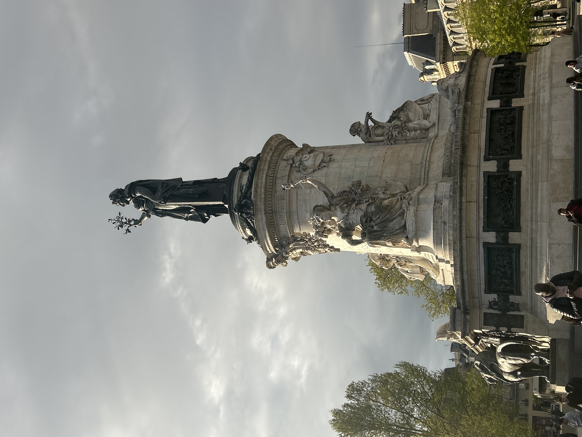 The image shows the Place de la République monument in Paris, France, a large central statue of Marianne (symbolizing the French Republic) atop a tall pedestal adorned with sculptures and bronze plaques. The sky is overcast. People are present in the foreground, seemingly walking or standing near the base of the monument. Trees with green foliage are visible, suggesting it is not winter. The surrounding buildings are typical Parisian architecture.