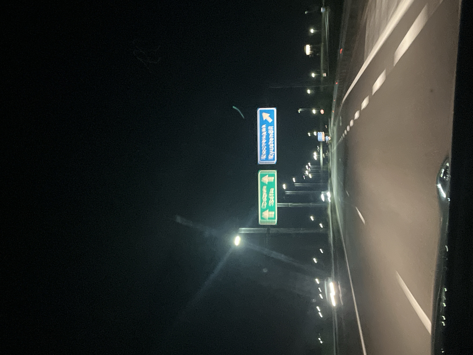 The image depicts a nighttime highway scene, likely taken from a vehicle. The highway stretches into the distance with white lane markings. Streetlights line the road. Road signs are visible indicating directions to specific locations: "Kostorhama" and "Kyustendil" are on one sign, while another sign displays locations with arrows pointing in different directions. The background is dark, suggesting it is nighttime. The scene appears to be in or near the city of Blajievo, Bulgaria.