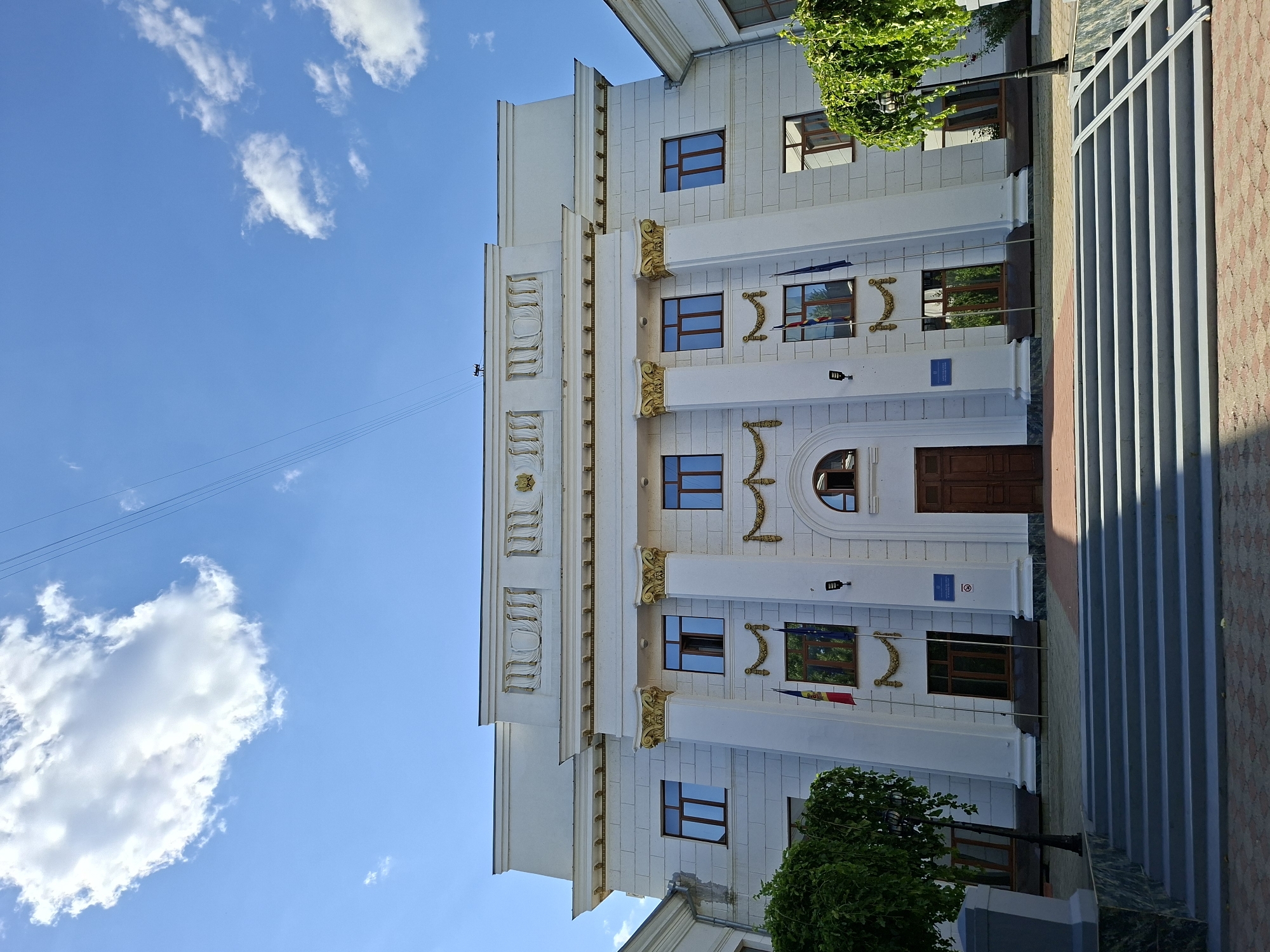 The image shows the main facade of the State University of Moldova, located in Chișinău, Moldova. The building is a large, white, neoclassical structure with a prominent central entrance and symmetrical wings. A wide set of grey stairs leads up to the entrance, flanked by manicured trees and lampposts. Two flags, one of Moldova and one of the European Union, are visible on poles in front of the building. The sky above is bright blue with a few scattered white clouds. The overall impression is one of a grand and well-maintained educational institution.