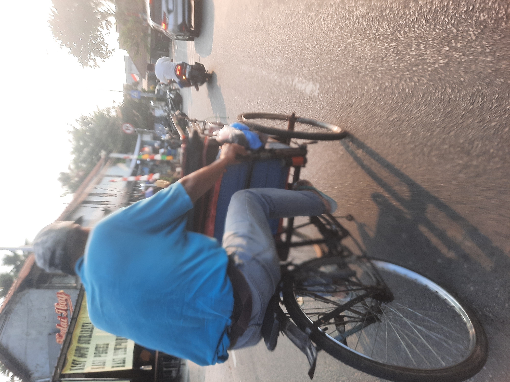 In North Jakarta, Indonesia, at approximately golden hour, a man wearing a blue t-shirt and a grey cap rides a pedal rickshaw. The rickshaw has a blue tarp covering its cargo area. The man is pedaling forward on a paved road, casting a shadow to his right. In the background, other vehicles are visible, including a silver car and a motorcycle with a rider. Buildings line the street on the left, with signage that appears to be for food establishments. The lighting suggests it is either sunrise or sunset, with a warm, hazy glow over the scene.