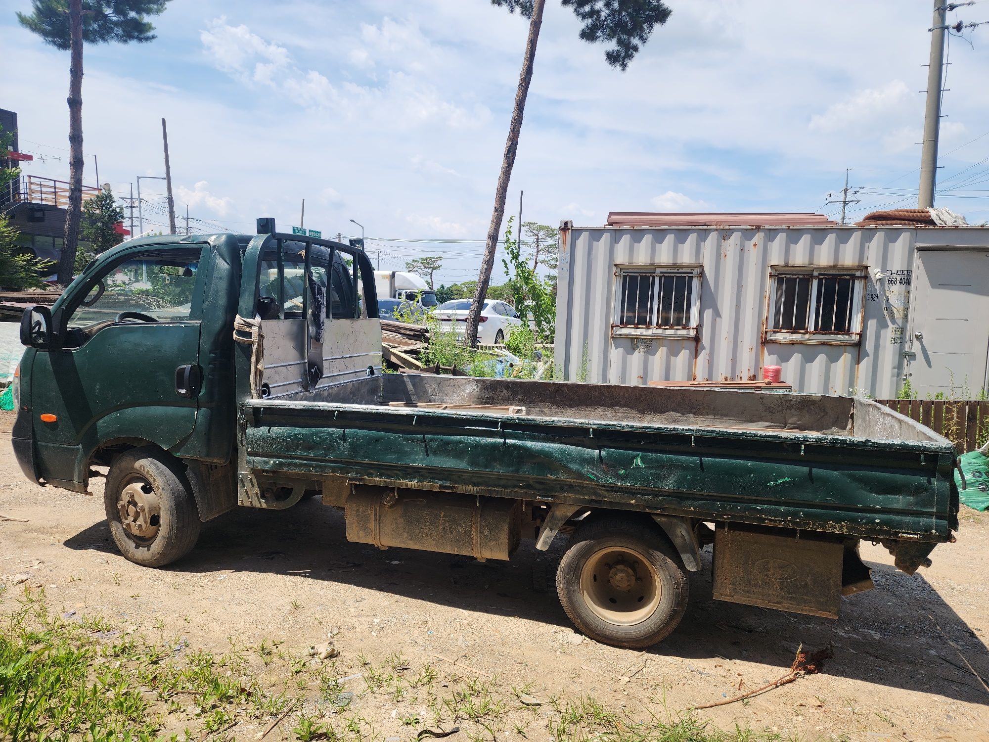 The image depicts a dark green flatbed truck parked on a dirt and gravel surface in Pyeongtaek-si, South Korea. The truck appears to be older and shows signs of wear. It has a metal bed that is empty, and the vehicle has a visible exhaust pipe and fuel tank. In the background, there is a large, weathered shipping container with barred windows, and beyond that, various structures including utility poles, power lines, and some vegetation. A tall, slender pine tree with sparse needles is prominent in the left half of the frame, reaching towards a partly cloudy blue sky. The scene suggests an outdoor storage or work area, possibly in a less developed part of the city. There are no people visible in the image.