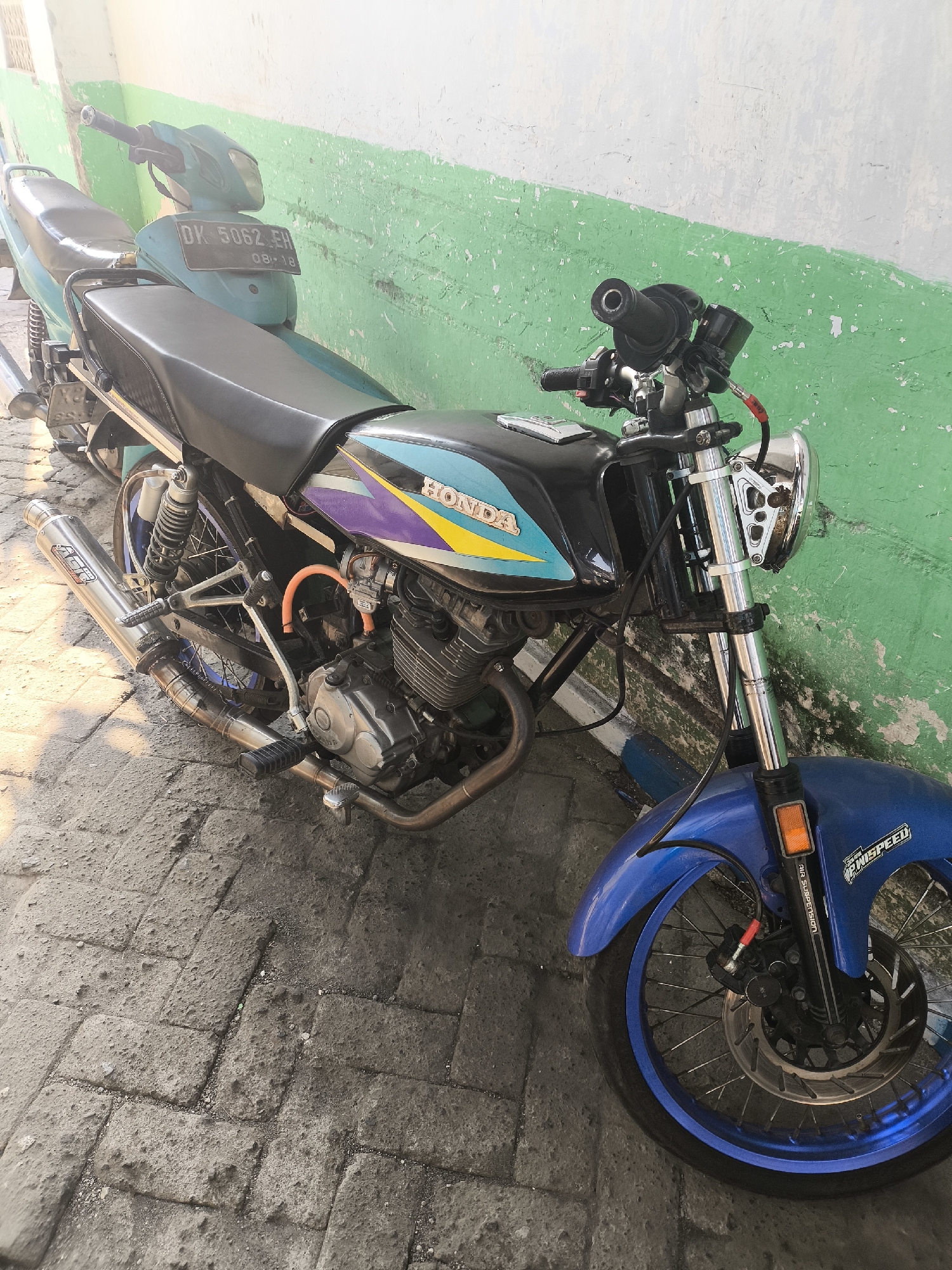 A blue and black Honda motorcycle is parked on a grey brick sidewalk in front of a green and white wall. The motorcycle has a black seat, a blue front fender, and a silver exhaust pipe. The license plate reads "DK 5062 EH 08-18". The wall is painted green on the bottom and white on the top, with some peeling paint. The sidewalk is made of interlocking grey bricks, with some dirt and debris scattered on it. The scene appears to be outdoors, in a city environment, possibly Lumajang, Indonesia, based on the license plate format. There are no people visible in the image.