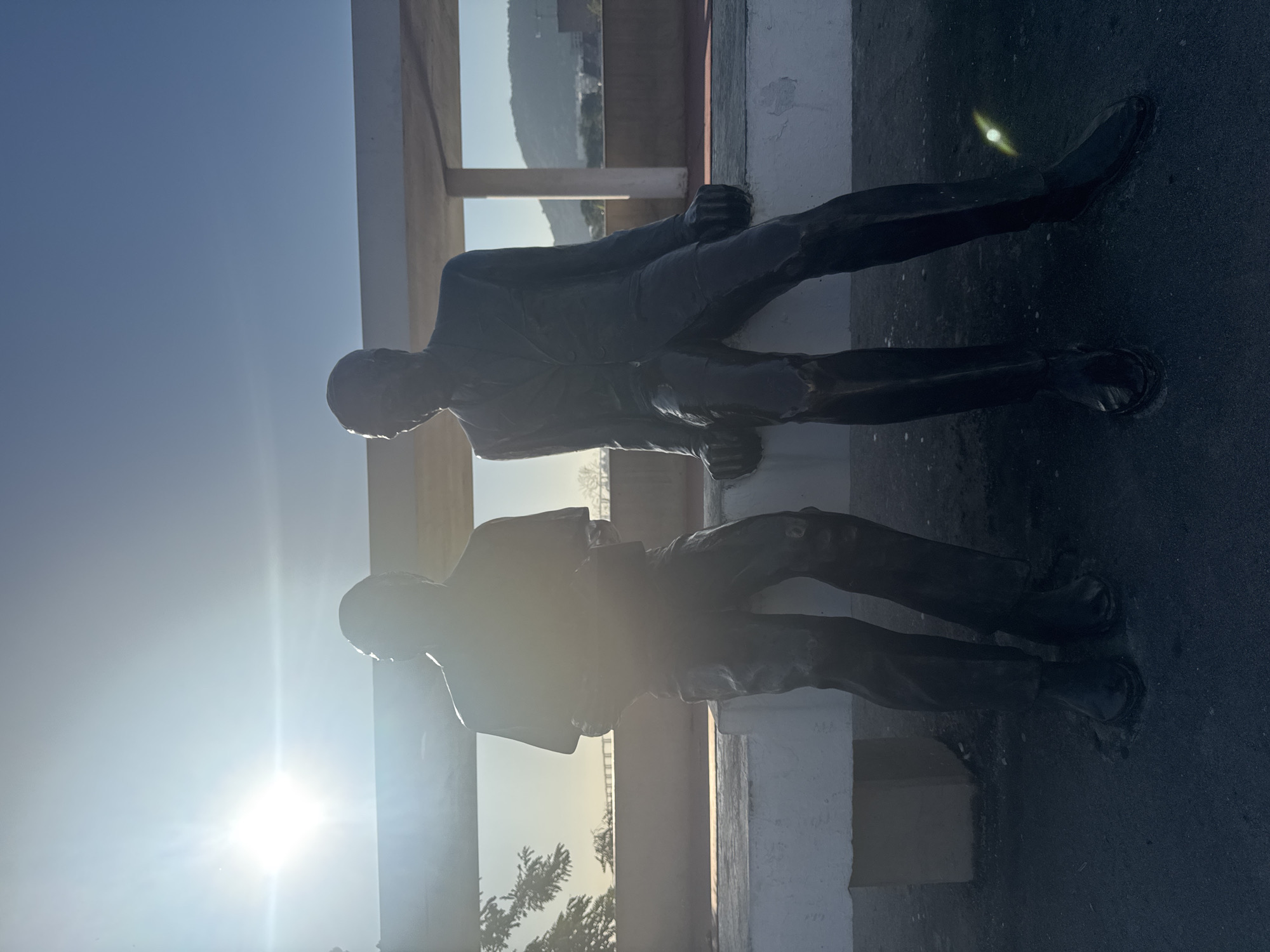 The image shows two life-size bronze statues of men sitting side-by-side on a white concrete bench. The statue on the left is wearing a collared shirt and pants, and is holding something in his hands. The statue on the right is wearing a suit and tie, with his hands resting on his knees. Both statues are facing to the right, with the statue on the right looking slightly towards the viewer. The statues are silhouetted against a bright sky with the sun shining intensely from the left, creating lens flare. In the background, there is a low-rise building with a white roof supported by pillars, and a hazy mountainous landscape beyond. This is the **Memorial Alfredo Sirkis** located in the **Parque da Cidade** in Niterói, Brazil, a public park offering panoramic views of the city, Guanabara Bay, and Rio de Janeiro. The lighting suggests it is either early morning or late afternoon.