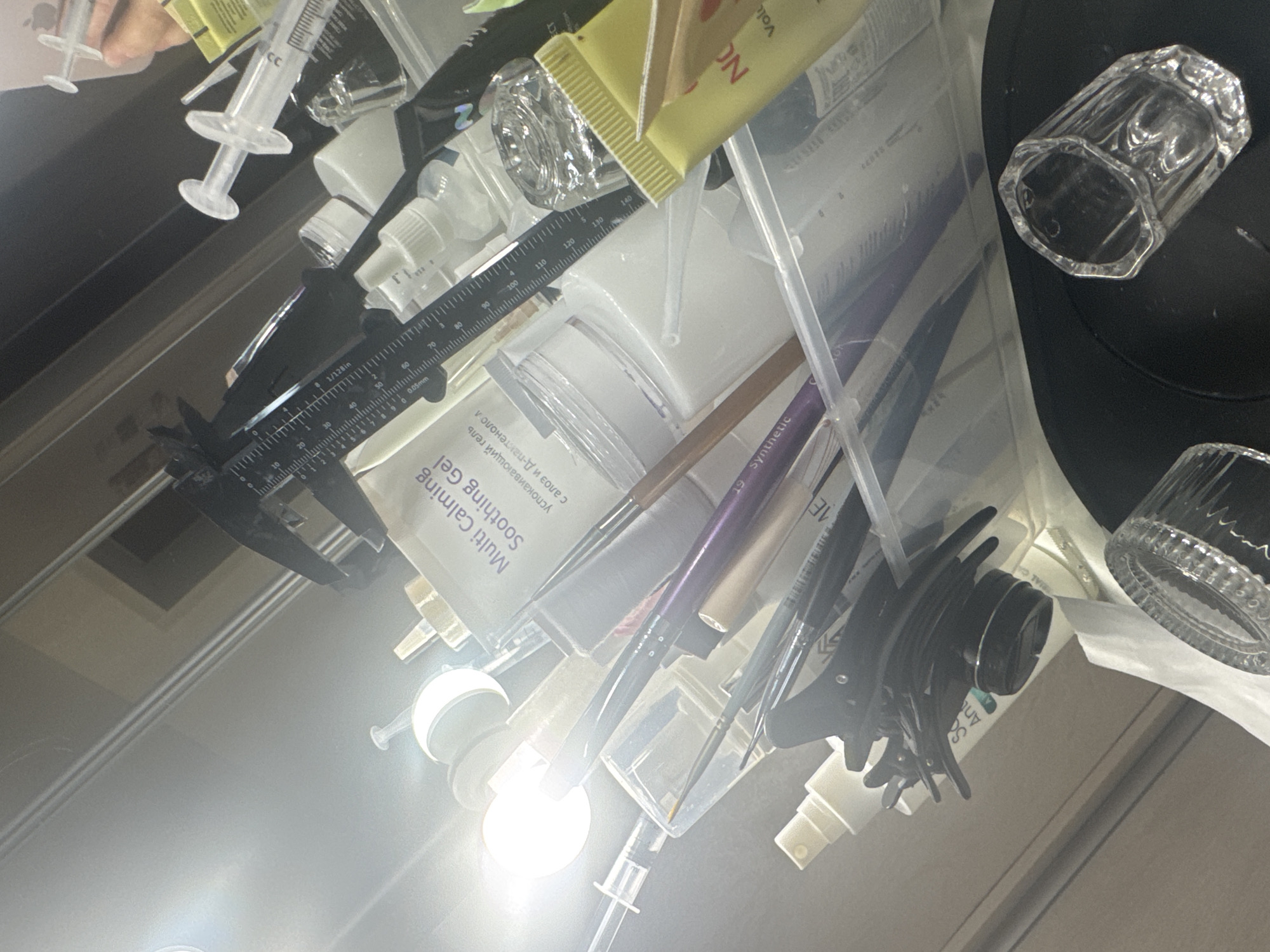 The image depicts a collection of beauty or cosmetic products, including syringes, bottles, tubes, brushes, and clips, organized within a transparent container. A ruler and caliper are also visible, suggesting precise measurements are being taken or utilized. The lighting appears to be artificial, with a bright light source creating glare in the upper left quadrant. The scene is likely a workspace or storage area for these items. There is no discernible information in the image to indicate a location in Saratov, Russia.