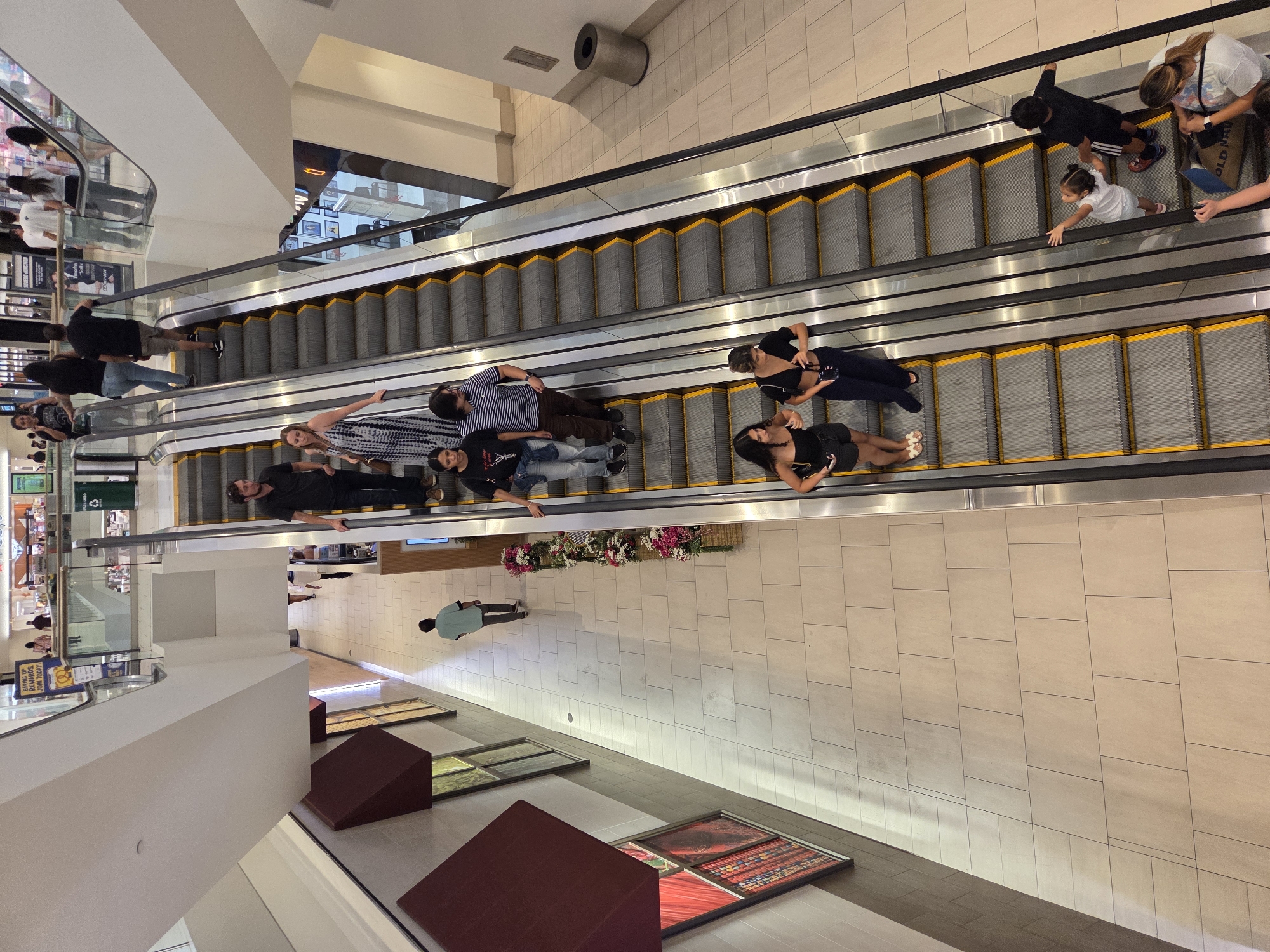 The image depicts an indoor escalator in a shopping mall in Burbank, California. Several people are visible on the escalators and in the surrounding area. On the left escalator, moving upwards, are three individuals: a man in a black shirt and dark pants, followed by a woman in a black and white patterned dress, and another woman in a black top and blue jeans. On the right escalator, also moving upwards, are a man in a black shirt and dark pants, a woman in a black top and dark pants, and two children. A young girl in a white shirt and shorts is holding onto the handrail, and a boy in a black t-shirt and shorts stands beside her. Further down the right escalator, a woman with blonde hair is holding a bag. In the background, there are shops and other shoppers visible. The mall features beige tiled flooring and white walls. There are also decorative planters with flowers along the right side of the walkway.