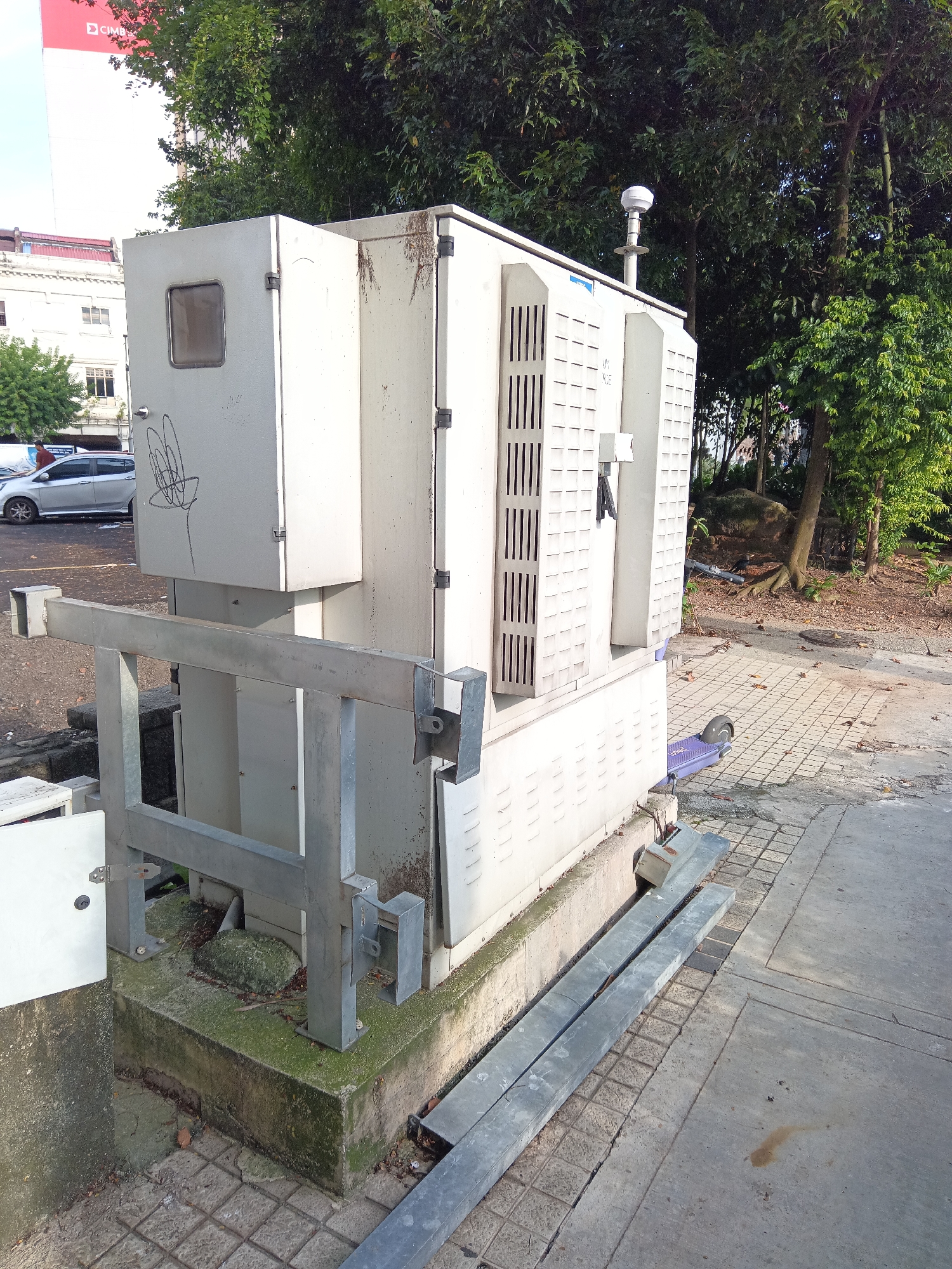This image shows a large, beige electrical box situated on a concrete platform in an outdoor urban environment. The box is rectangular with various panels, vents, and a small window on its left side. A metal railing with a silver finish is attached to the front of the platform. The area surrounding the platform is paved with square tiles and concrete. Several metal beams are placed on the ground next to the railing. In the background, there are trees with green foliage, a building with a red and white advertisement, and a silver car. A purple electric scooter is visible on the tiled ground to the right of the electrical box. The scene appears to be located in a park or public space in Kuala Lumpur, Malaysia, with evidence of some wear and tear on the electrical box and surrounding structures.