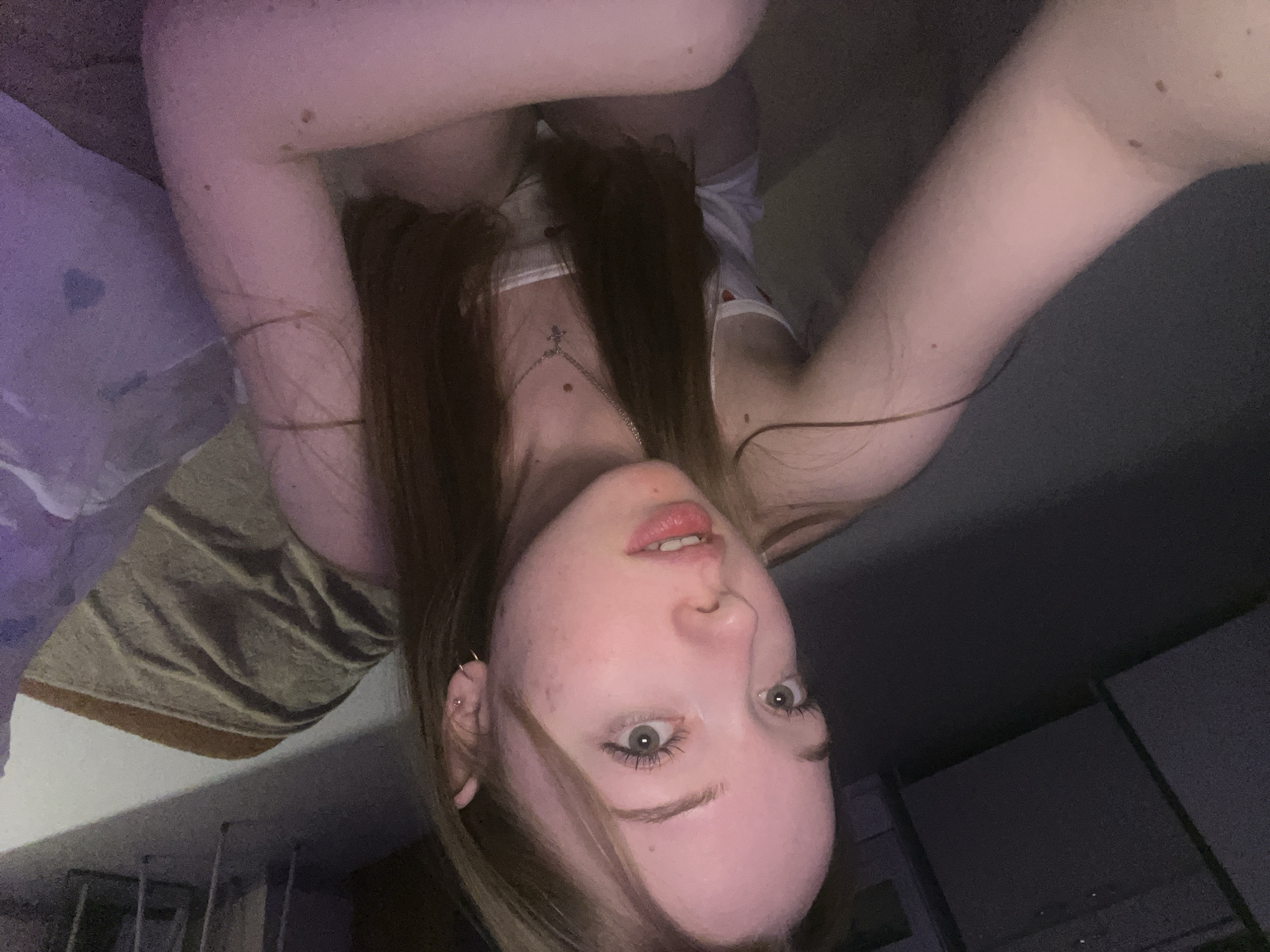 A portrait-oriented selfie captures a young female from an upside-down perspective, suggesting she is lying down. Her face, positioned in the lower center of the frame, looks directly at the camera with a neutral yet direct gaze. She has long, dark brown hair that frames her face and spreads around her, fair skin, and prominent light-colored eyes (likely green or hazel) with visible mascara. Her lips are slightly parted, revealing her upper teeth.
She is wearing a light-colored (possibly white) camisole or tank top. A silver-toned necklace with a cross pendant hangs on her chest. Small hoop earrings are visible in her left ear. Her arms are raised and partially visible on either side of her head, showing fair skin with a few moles.
The setting appears to be indoors, characterized by dim lighting consistent with evening or nighttime. In the upper left background, a piece of textured fabric, possibly a blanket or towel, is visible. The remainder of the background is dark and indistinct, with faint outlines of what might be furniture or architectural features in the lower right. The overall impression is an intimate and casual moment. There is no visible text in the image. The image originates from Chaikovsky, Russia.