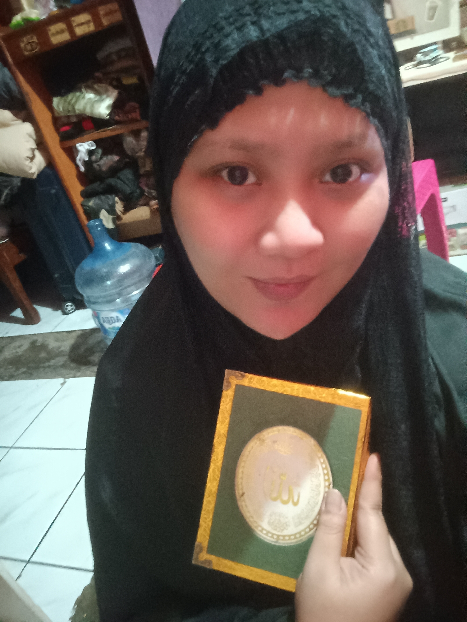 A medium shot captures a young woman wearing a black hijab, looking directly at the camera. She is holding a small, square, gold-trimmed green box in her right hand. The box features a circular metallic design with Arabic script. The background is cluttered, with a wooden shelf unit filled with various items, a large blue water dispenser bottle, and a pink stool visible. The flooring appears to be tiled. The image is taken indoors, suggesting a residential setting. The lighting is somewhat dim, with a warm cast over the woman's face.