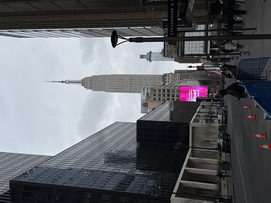 A street scene in New York City features the Empire State Building in the distance, partially obscured by buildings. A large glass skyscraper dominates the foreground. Traffic cones line the street, and pedestrian barriers are visible. A bright pink digital billboard displays an advertisement. Street signs for steakhouses and other businesses are present. The sky is overcast. People walk along the sidewalks.
