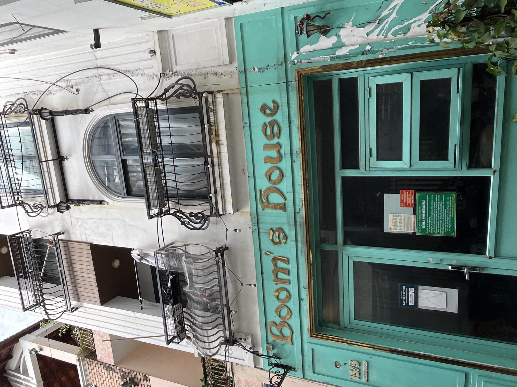 This image captures the facade of a multi-story building in Đà Nẵng, Vietnam, under bright daylight. The ground floor features a distinctive teal-colored front with a large glass door and windows, adorned with gold, embossed lettering spelling "Boom's House." Below this, "15/5" and "LE HONG PHONG" are visible, indicating the address. An old-fashioned, ornate lantern is mounted above the entrance.
The upper floors are painted in a lighter, off-white or light gray, showing signs of wear with peeling paint and some cracks. These floors are characterized by decorative wrought iron balconies and window grates, some of which appear to be protecting air conditioning units or storage areas. One notable window on the second floor is semi-circular or arched, framed by similar wrought iron work. Further up, additional balconies with modern wooden paneling and smaller round windows can be seen.
On the glass door of "Boom's House," several signs are affixed. One prominent green sign reads "TỔ DÂN PHỐ KHÔNG RÁC" (Residential Area No Trash). Another sign, mostly obscured, states "CĂN HỘ CHO THUẾ" (Apartment for Rent), with a QR code and a phone number partly visible. The overall scene suggests a vibrant urban environment with a mix of commercial and residential spaces, reflecting the local architecture of Đà Nẵng. No people are visible, and the weather appears clear and sunny.