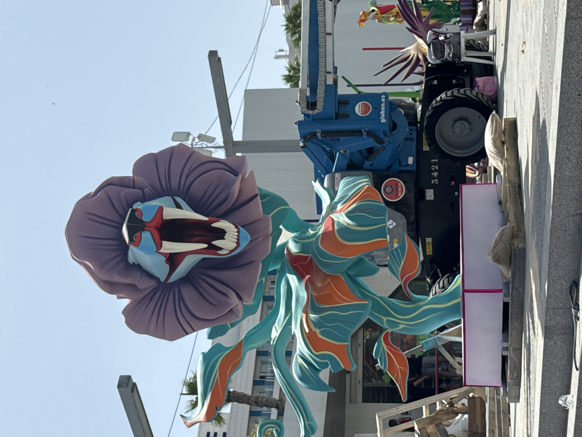 An outdoor scene shows a large, colorful sculpture resembling a monstrous flower with a lion's head at its center, in Unknown, Unknown. The sculpture is predominantly purple, teal, and orange, with the lion's head featuring a gaping mouth, sharp teeth, and blue and red accents around its eyes and snout. The sculpture is supported by a teal, organic-shaped structure that extends downwards.
Behind the sculpture, a bright blue aerial work platform is parked, with the brand name "globien.es" visible on its side. The platform has a large wheel and part of its mechanical arm is extended upwards. In the background, several white buildings with windows are visible, along with palm trees and utility poles with wires. The sky is clear and blue.
In the foreground, concrete steps lead to a paved area where the sculpture is situated. A pink-topped, rectangular platform is placed on wooden pallets. A burlap sack rests on some wooden planks near the platform. To the right of the aerial work platform, a white plastic chair is positioned, with a backpack on it. Other colorful elements of what appear to be more sculptures or decorations are partially visible on the right side of the image. A few individuals are visible in the background, seemingly observing or working on the structures.